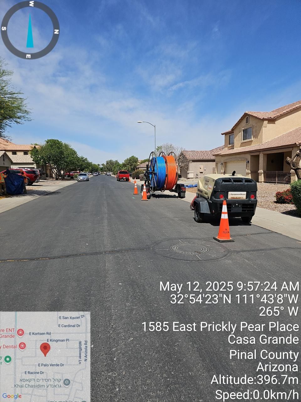Street view with parked vehicles, orange cones, and a large spool on a residential road under a blue sky