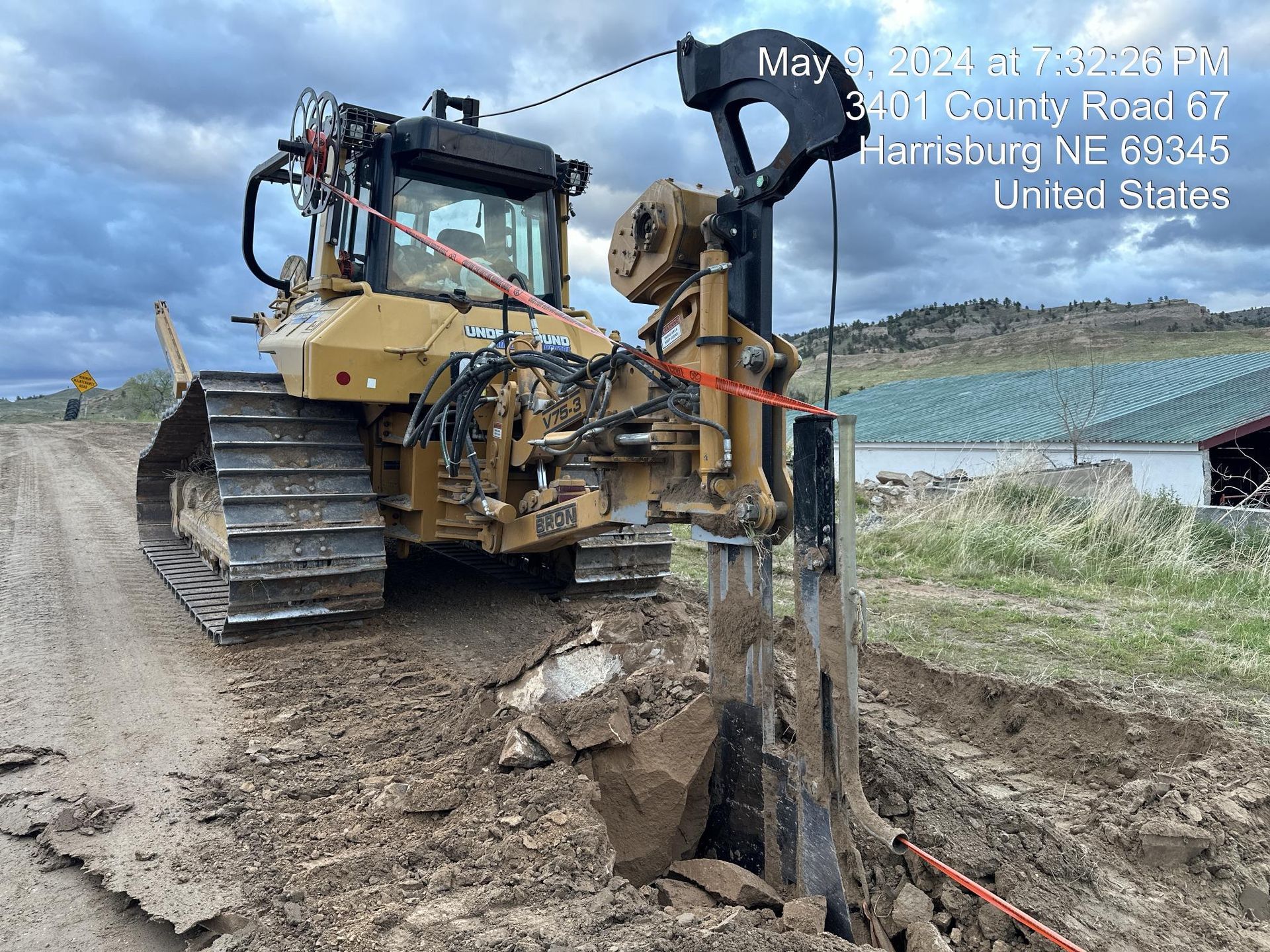 Bulldozer trenching muddy roadside beside a lake, with a utility pole and power lines nearby