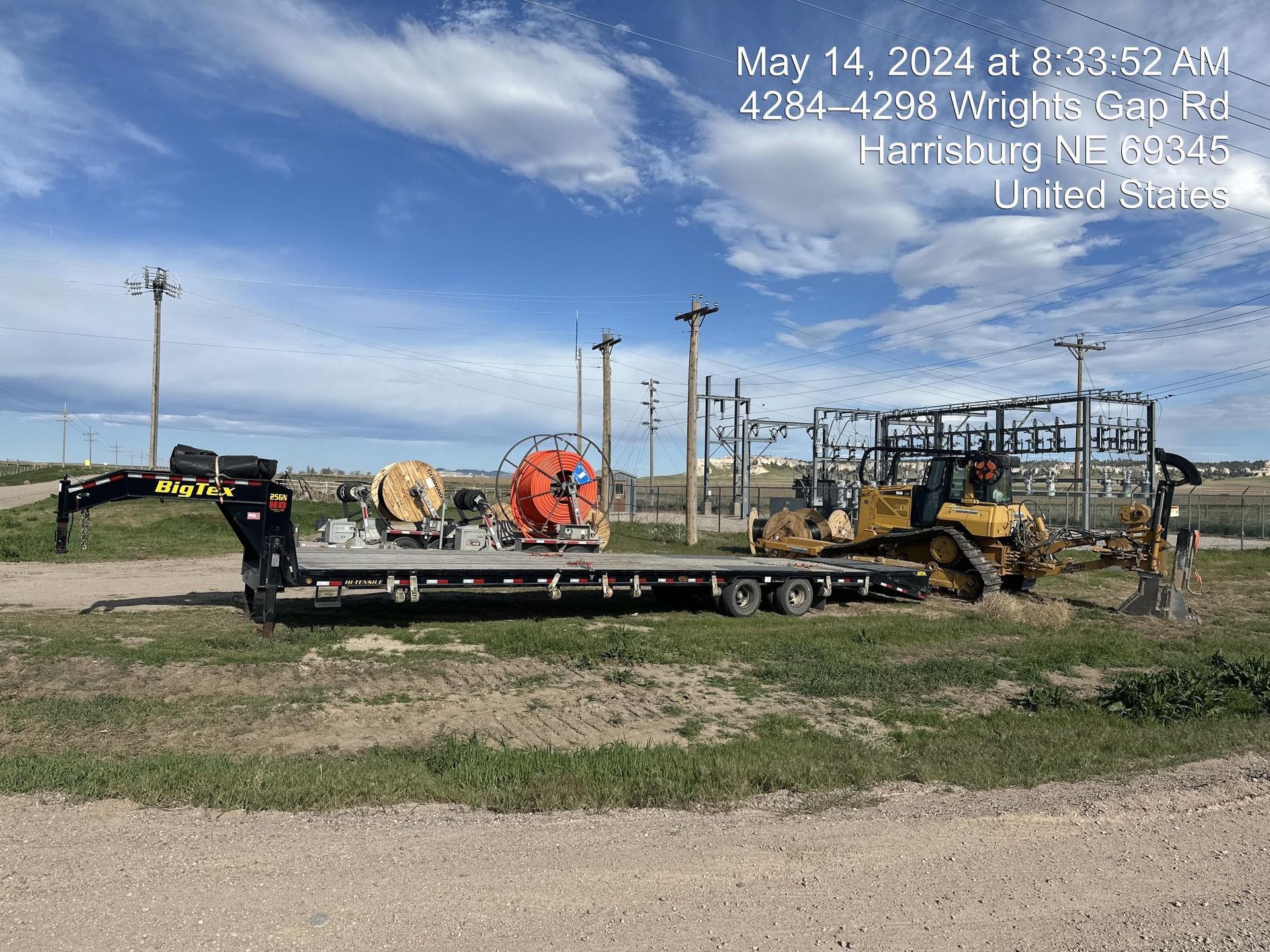 Flatbed trailer with construction equipment parked on a dirt lot under a partly cloudy sky.