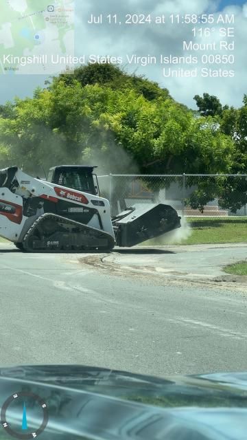 Skid-steer loader on a suburban street plows through a fence near a tree, with water spraying nearby.