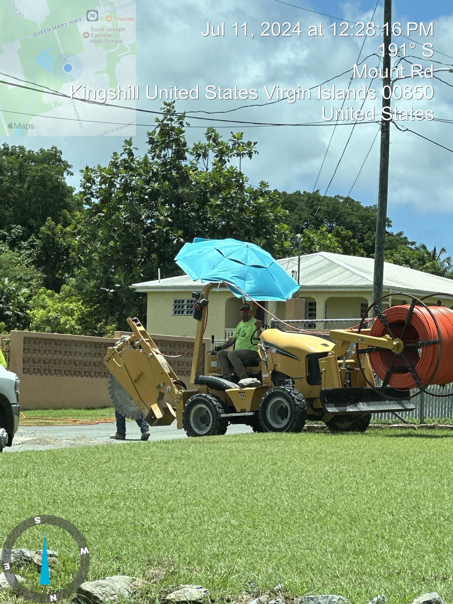 Yellow street-cleaning vehicle with blue umbrella parked on a grassy roadside near a brick wall and house.