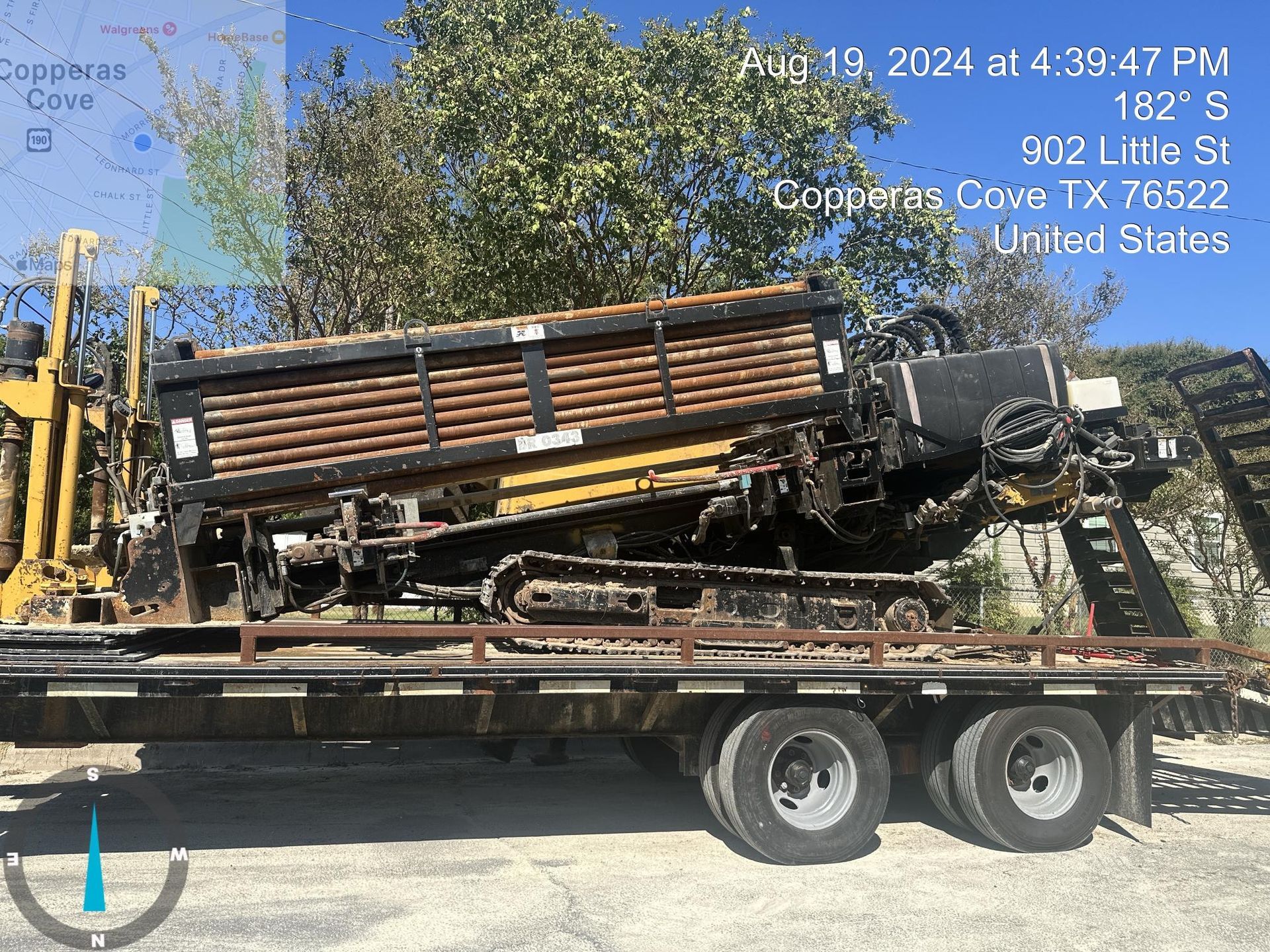 Damaged yellow tractor loaded on a flatbed trailer under trees, with wrecked front end visible