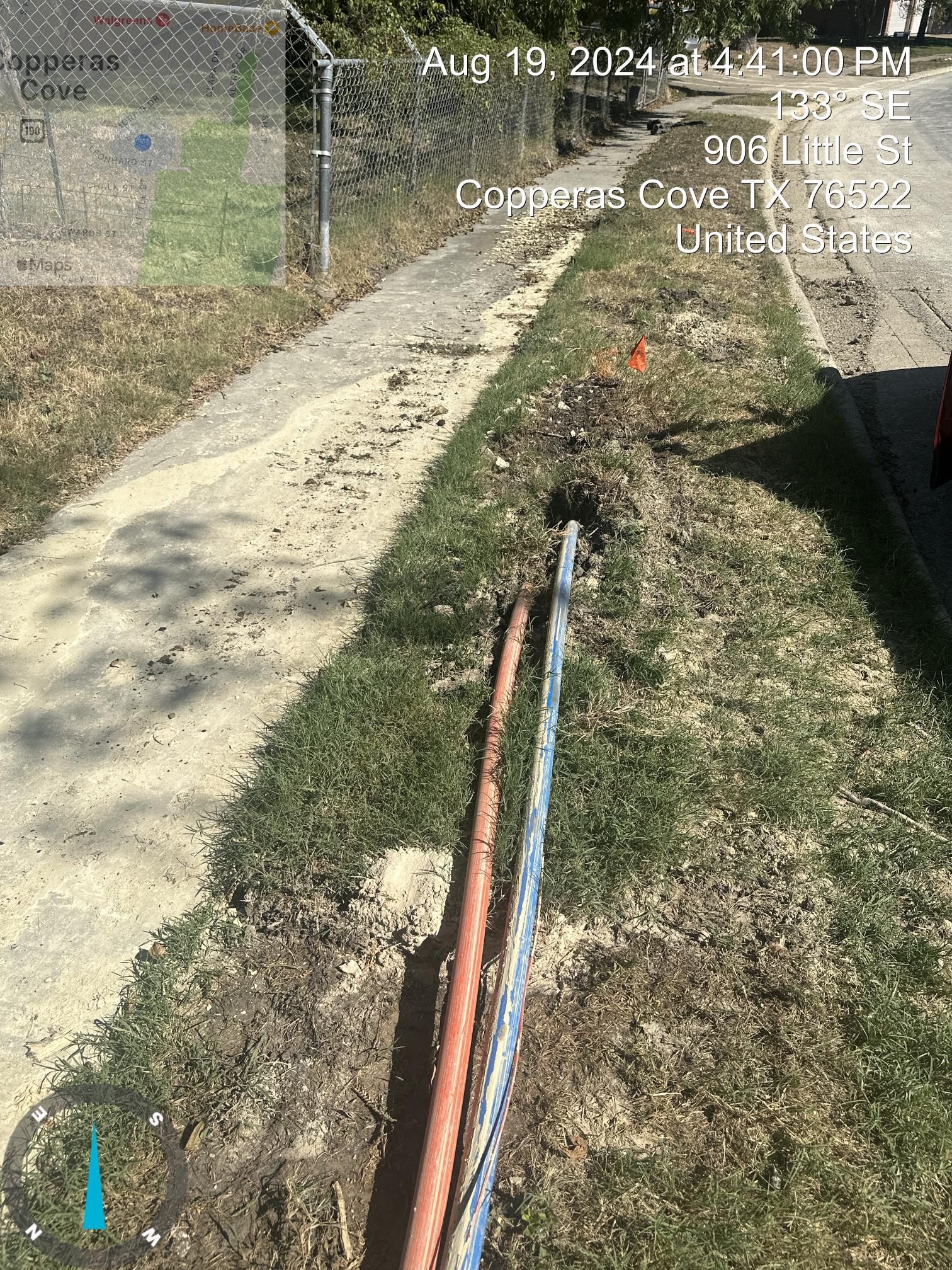 Sidewalk with exposed utility pipes running along a grassy edge beside a fence and road.