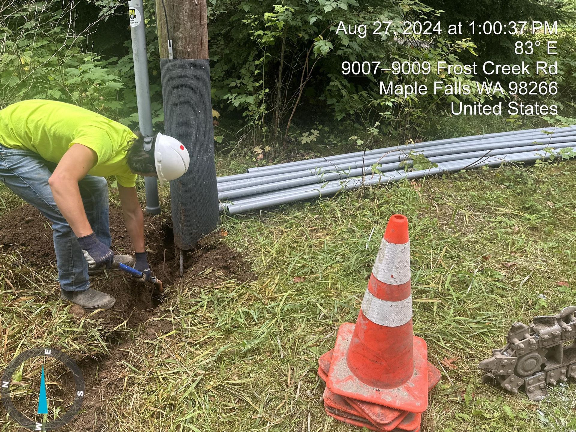 Worker in neon shirt and hard hat digs around a utility pole beside orange cones and stacked pipes.