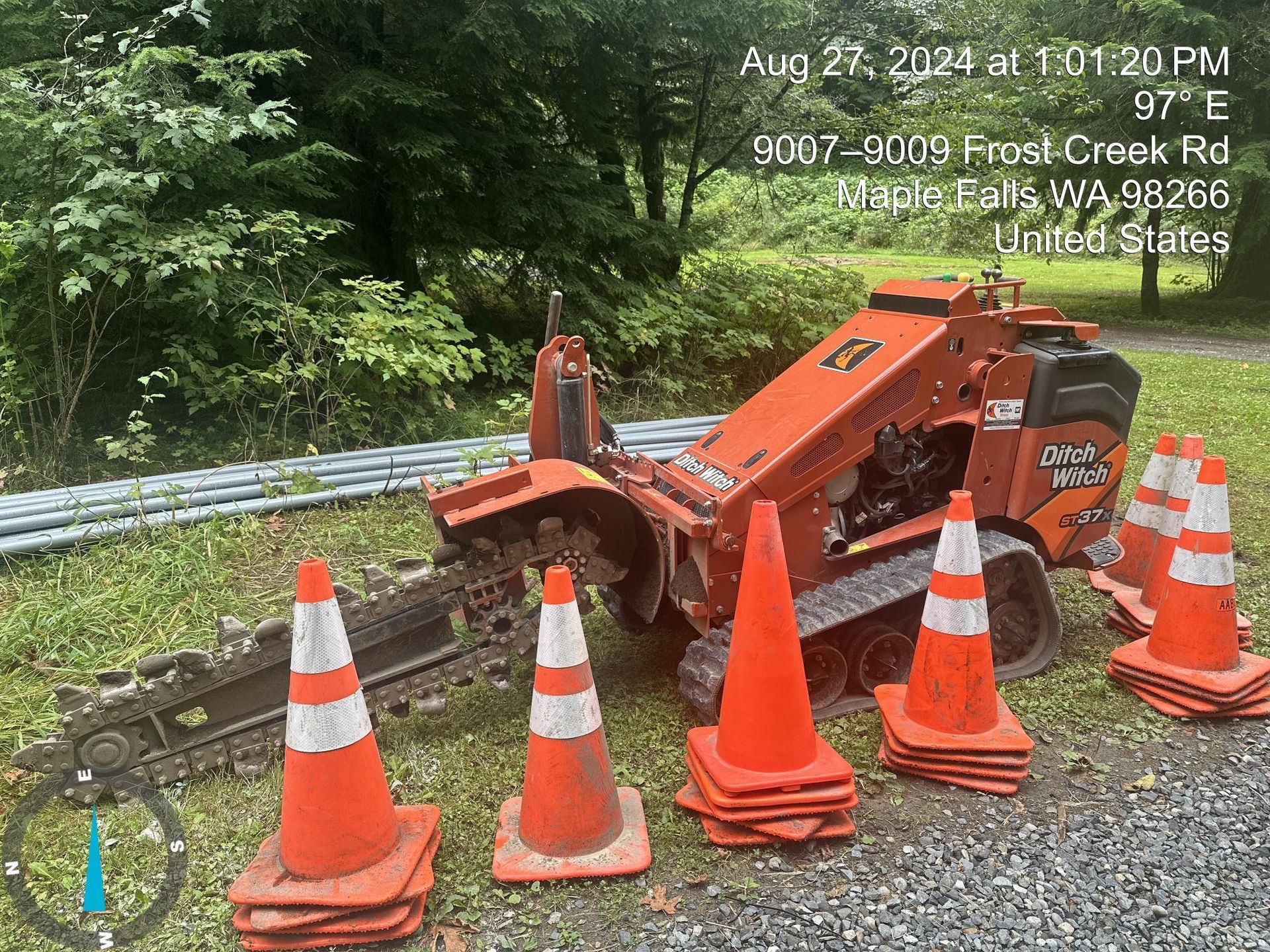 Orange road work machine and traffic cones blocking a gravel roadside lane near a guardrail.