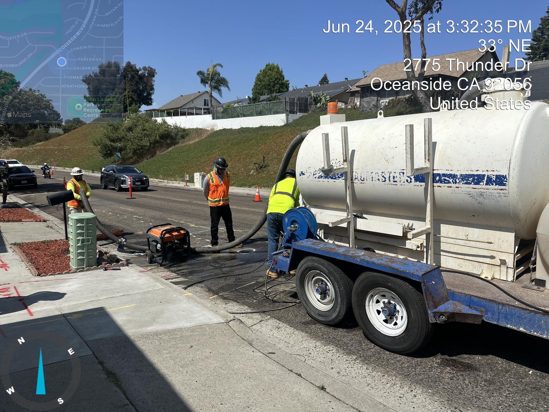 Road crew repairing a cracked street beside a white tanker truck and orange cones on a sunny hillside road