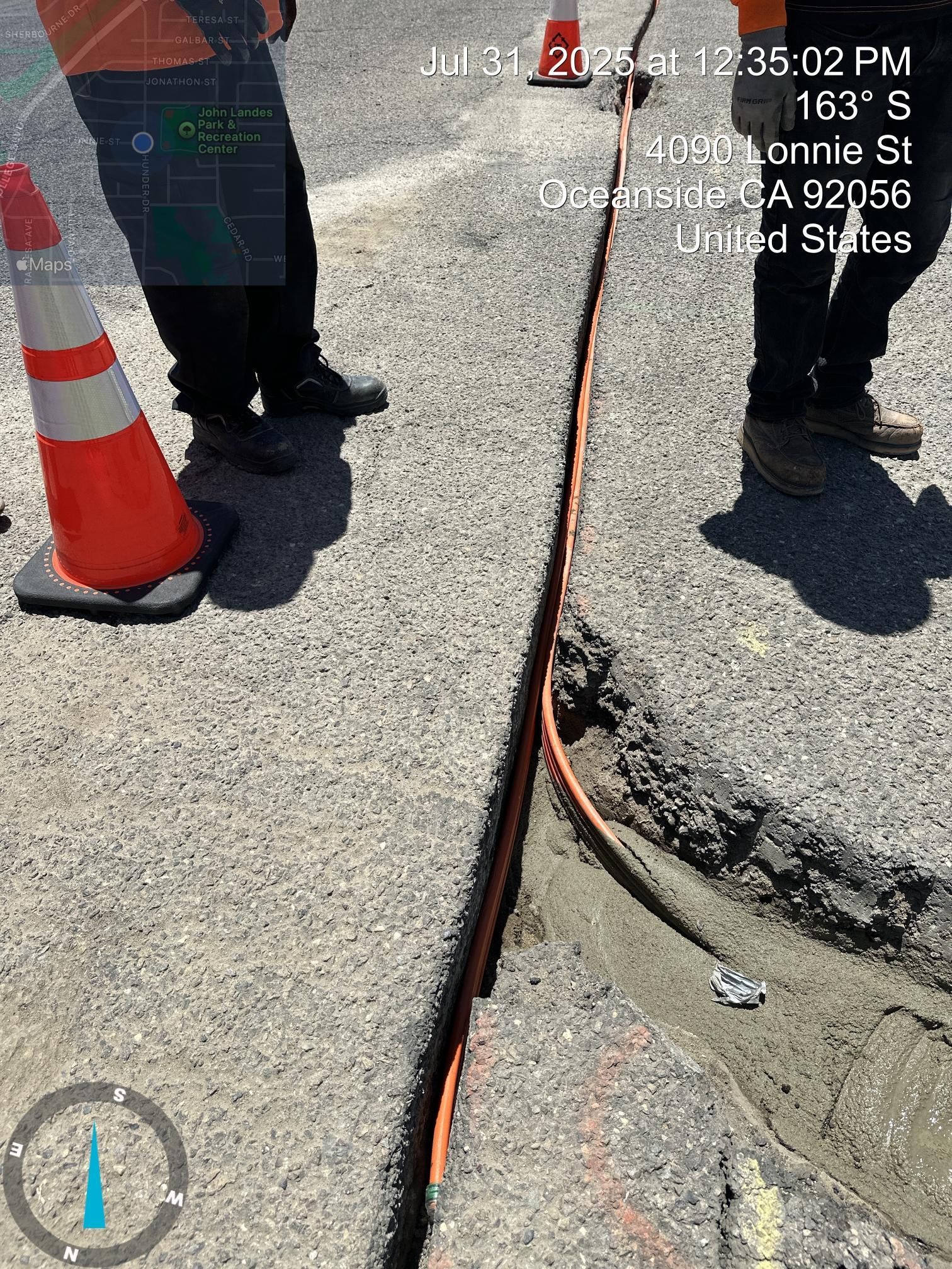 Orange traffic cone and workers’ feet beside a cracked sidewalk with an exposed cable.