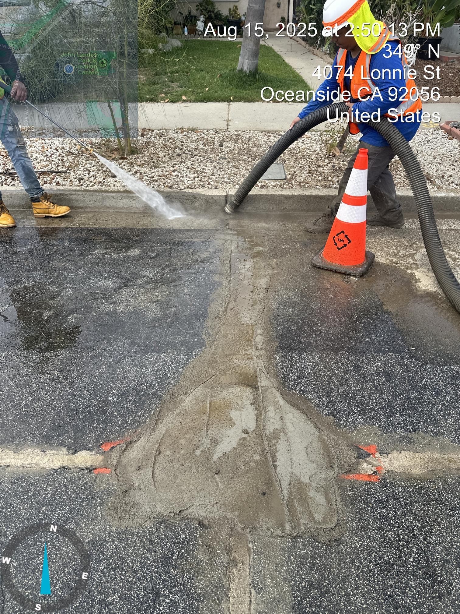 Construction worker spraying water over a wet asphalt patch beside orange cones and hoses on a street
