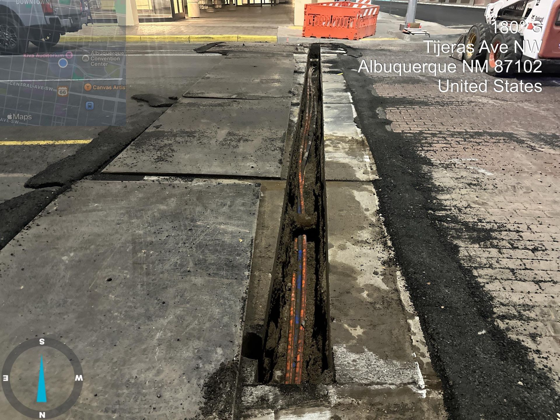 Roadwork trench cut through worn asphalt beside a curb and orange construction barrier.