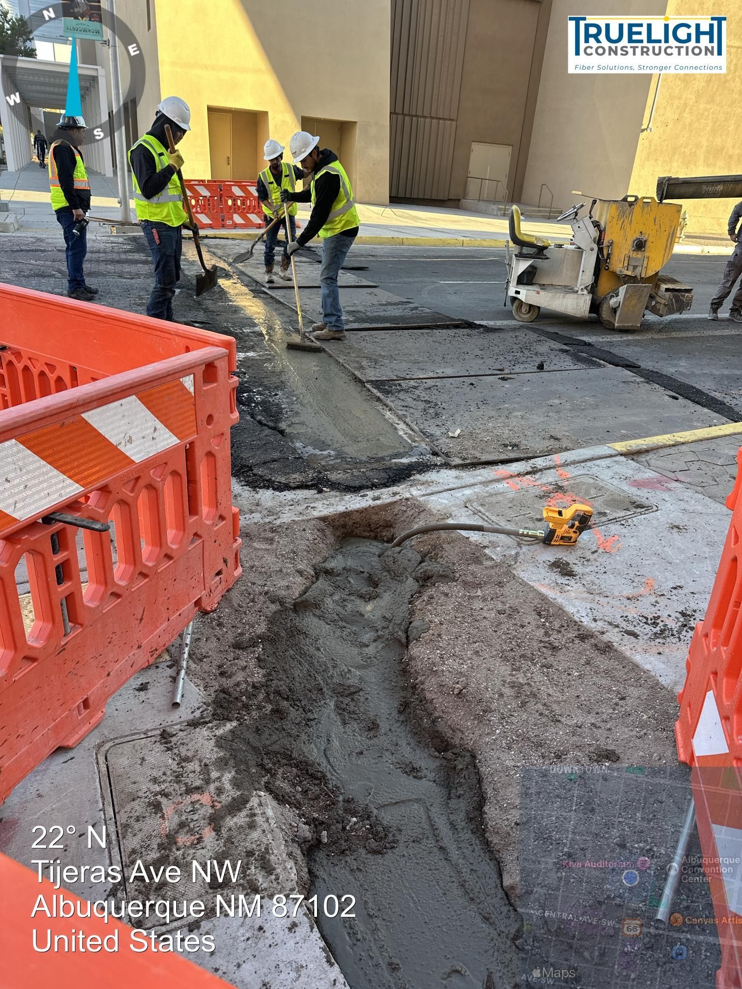 Construction crew pouring concrete into a road trench beside orange barriers and a yellow machine