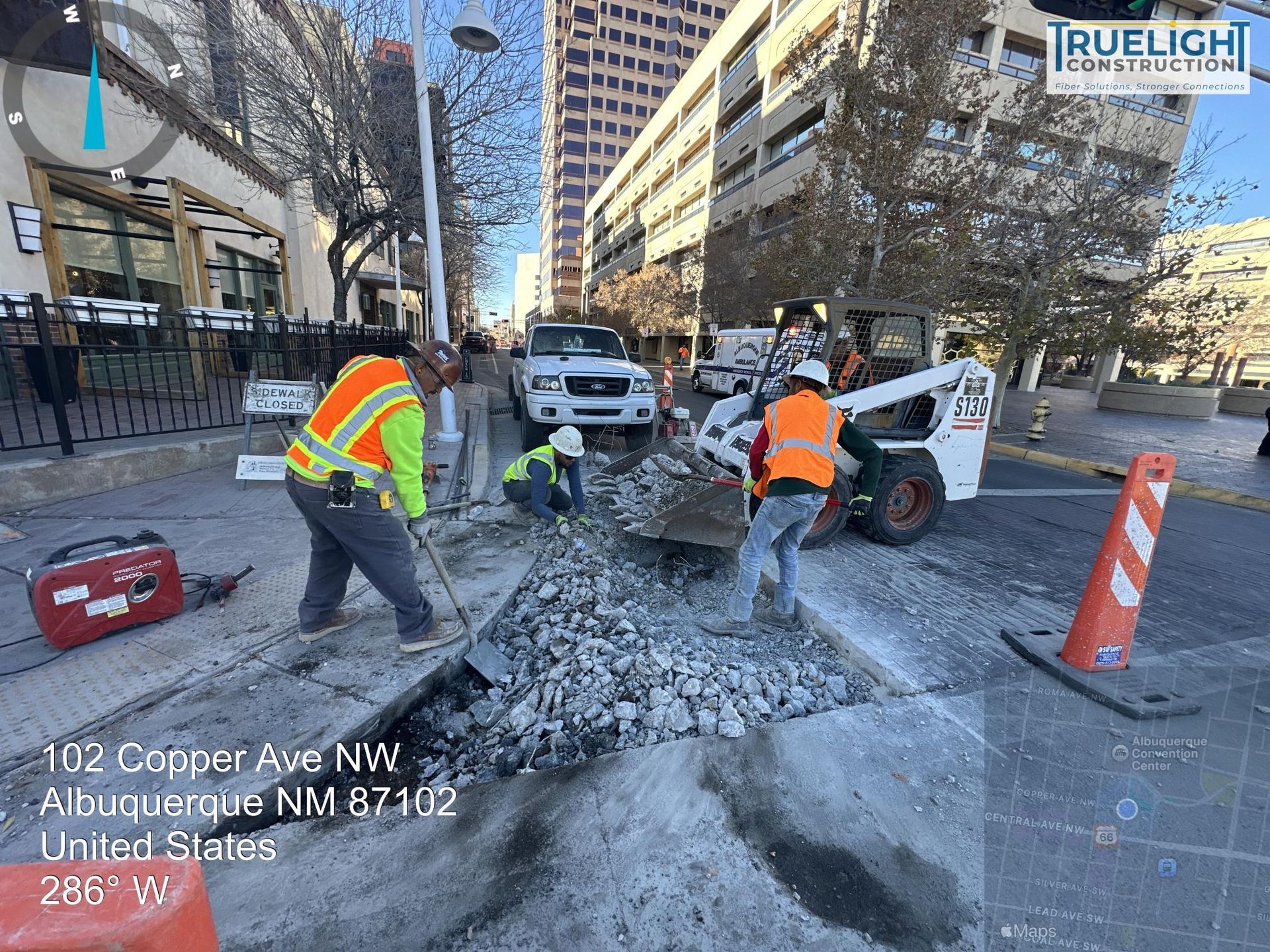 Workers in orange vests repair a broken street at Copper Ave NW with a skid steer and traffic cones