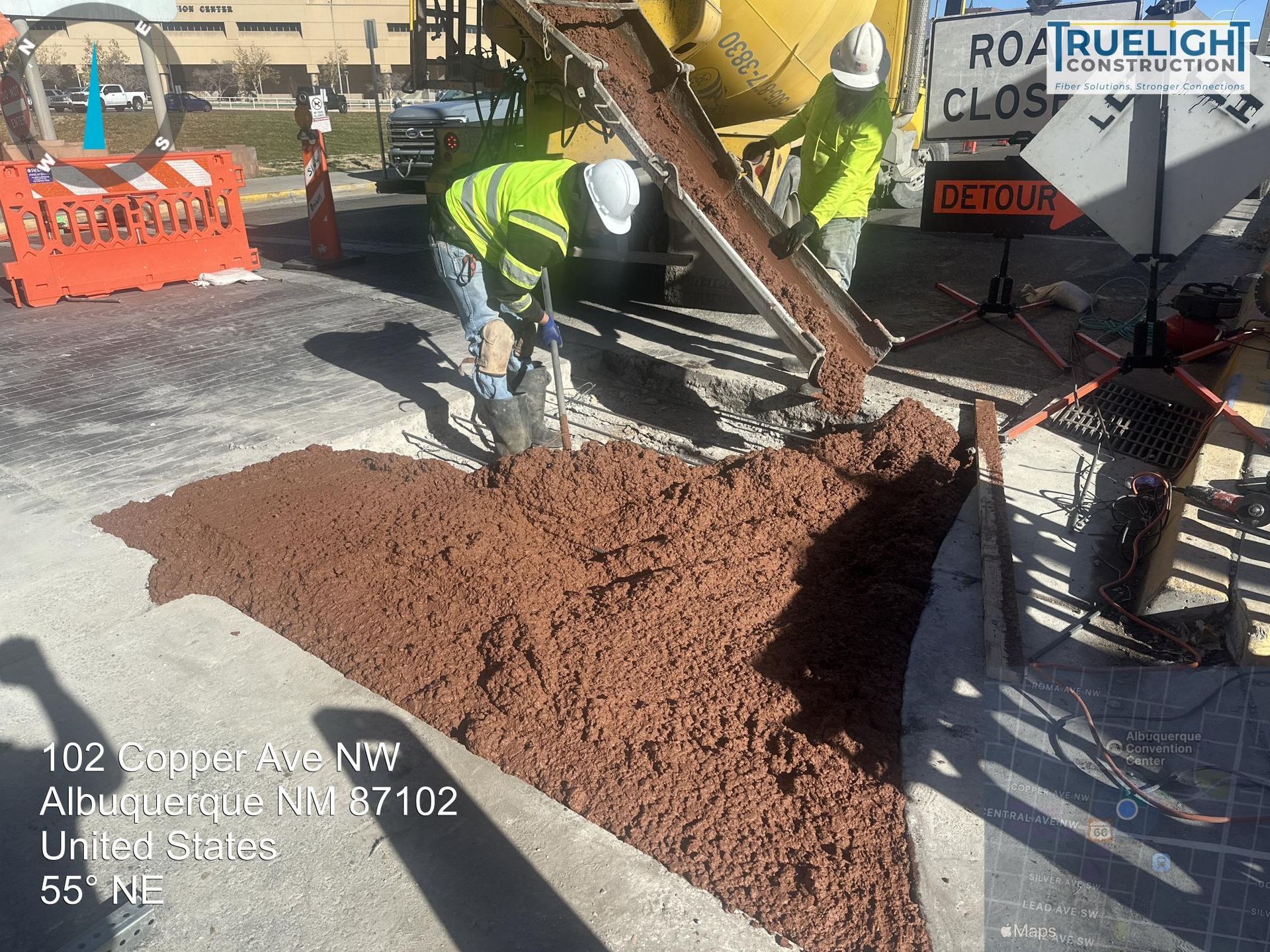 Workers pour red gravel from a cement mixer at a road construction site.