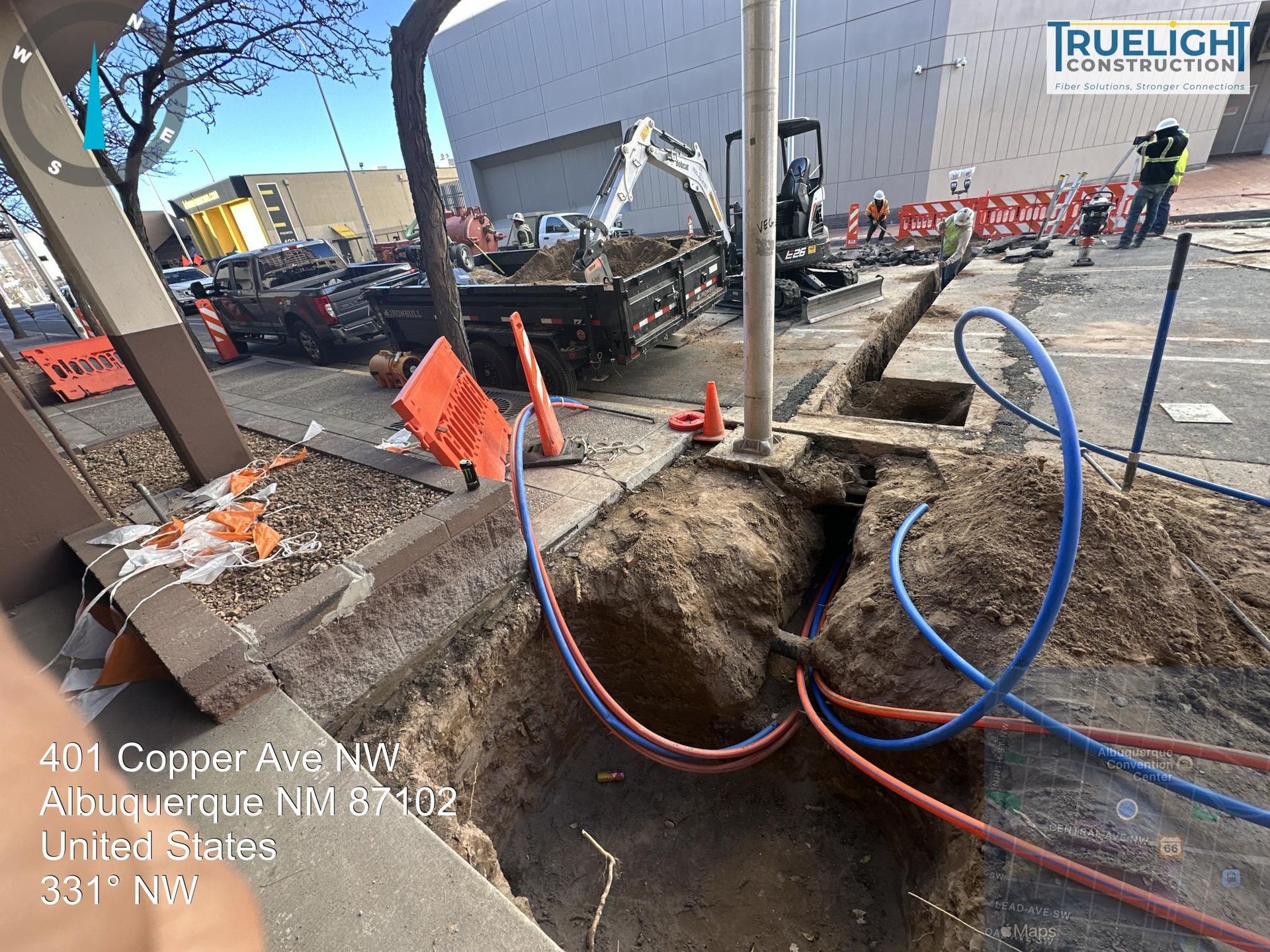 Road excavation with exposed utility lines, orange cones, and blue/red hoses at a construction site
