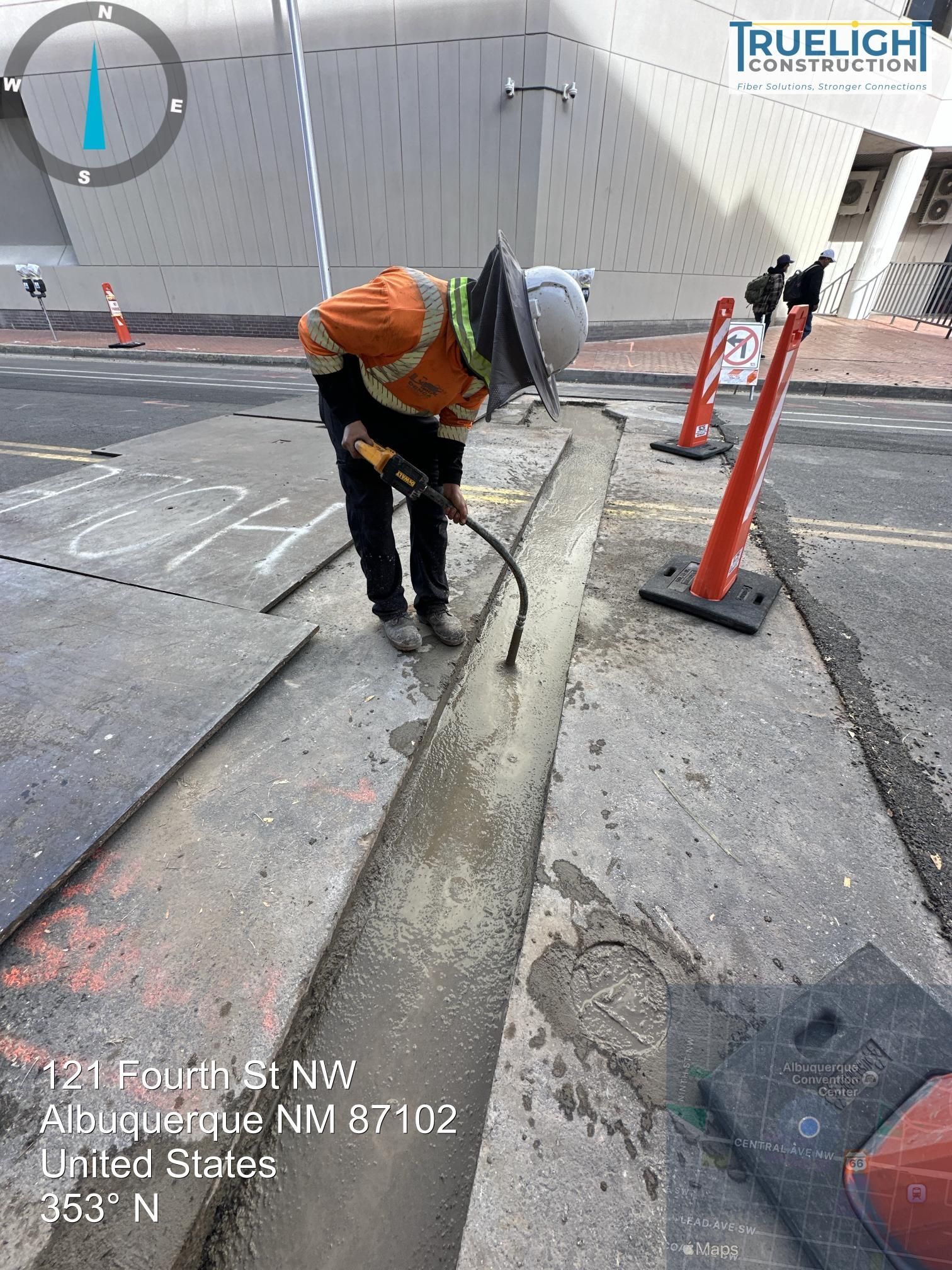 Worker in orange safety vest and hard hat using a jackhammer on a sidewalk near cones and a curb ramp