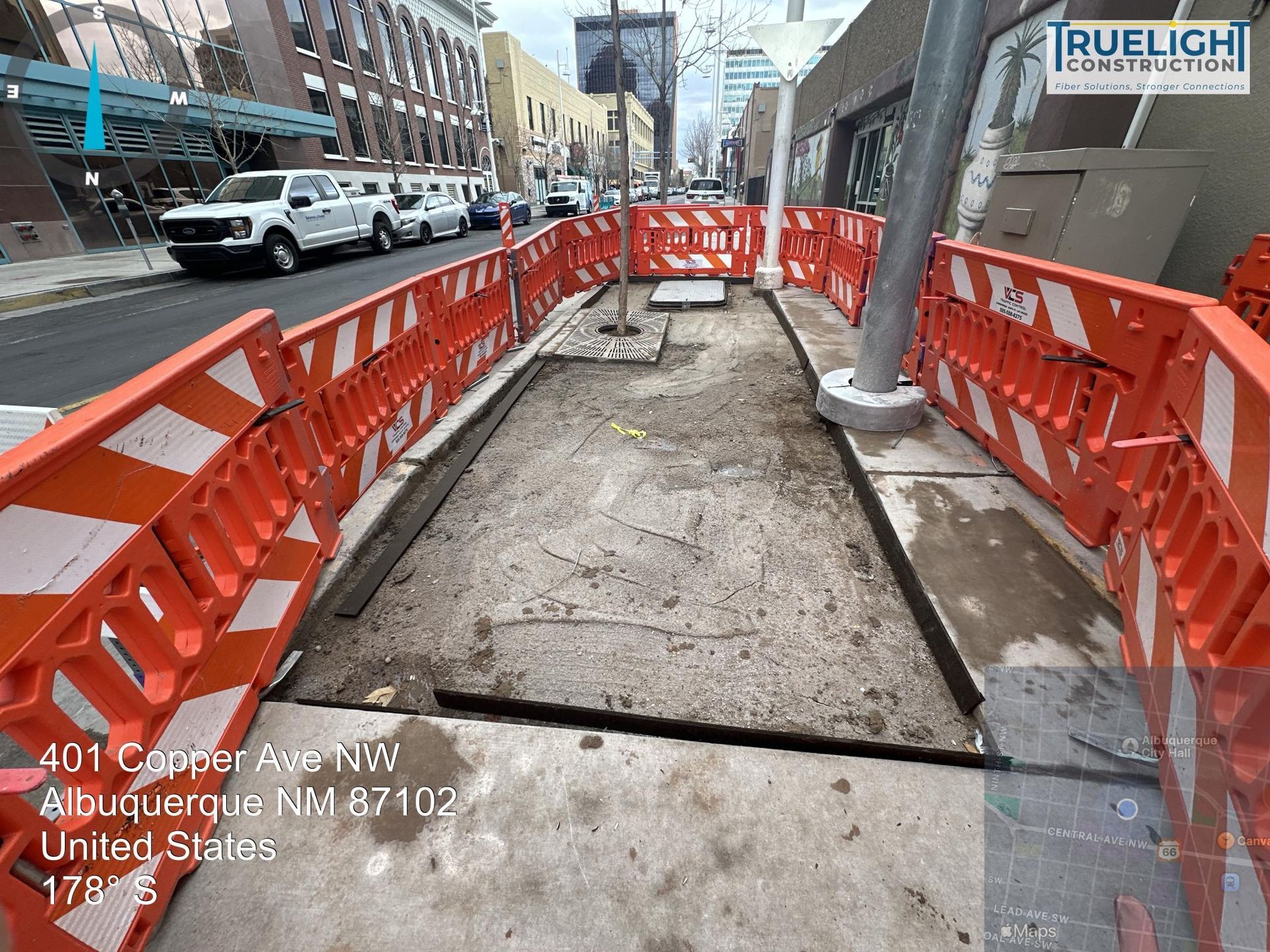 Orange barricades border a damaged sidewalk trench along a city street, near parked cars and buildings.