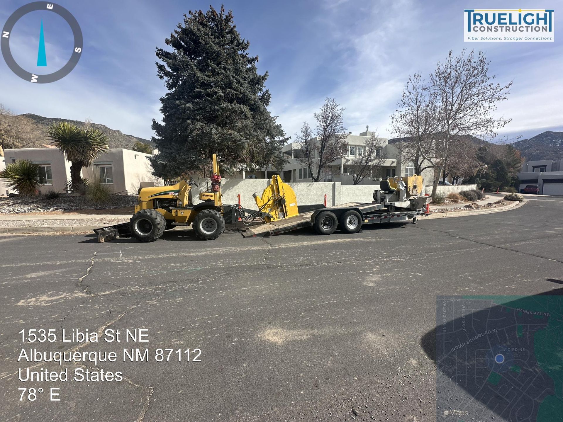 Yellow tractor towing equipment on a flatbed trailer in a residential street with mountains in the background