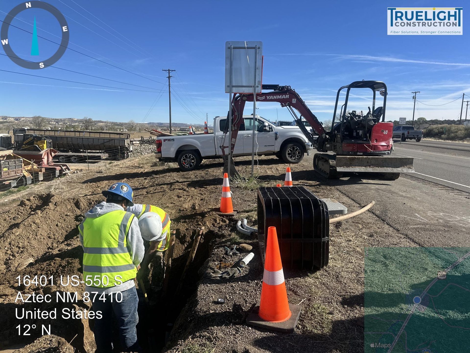 Utility worker by roadside trench with excavator, truck, cones, and survey equipment under clear blue sky