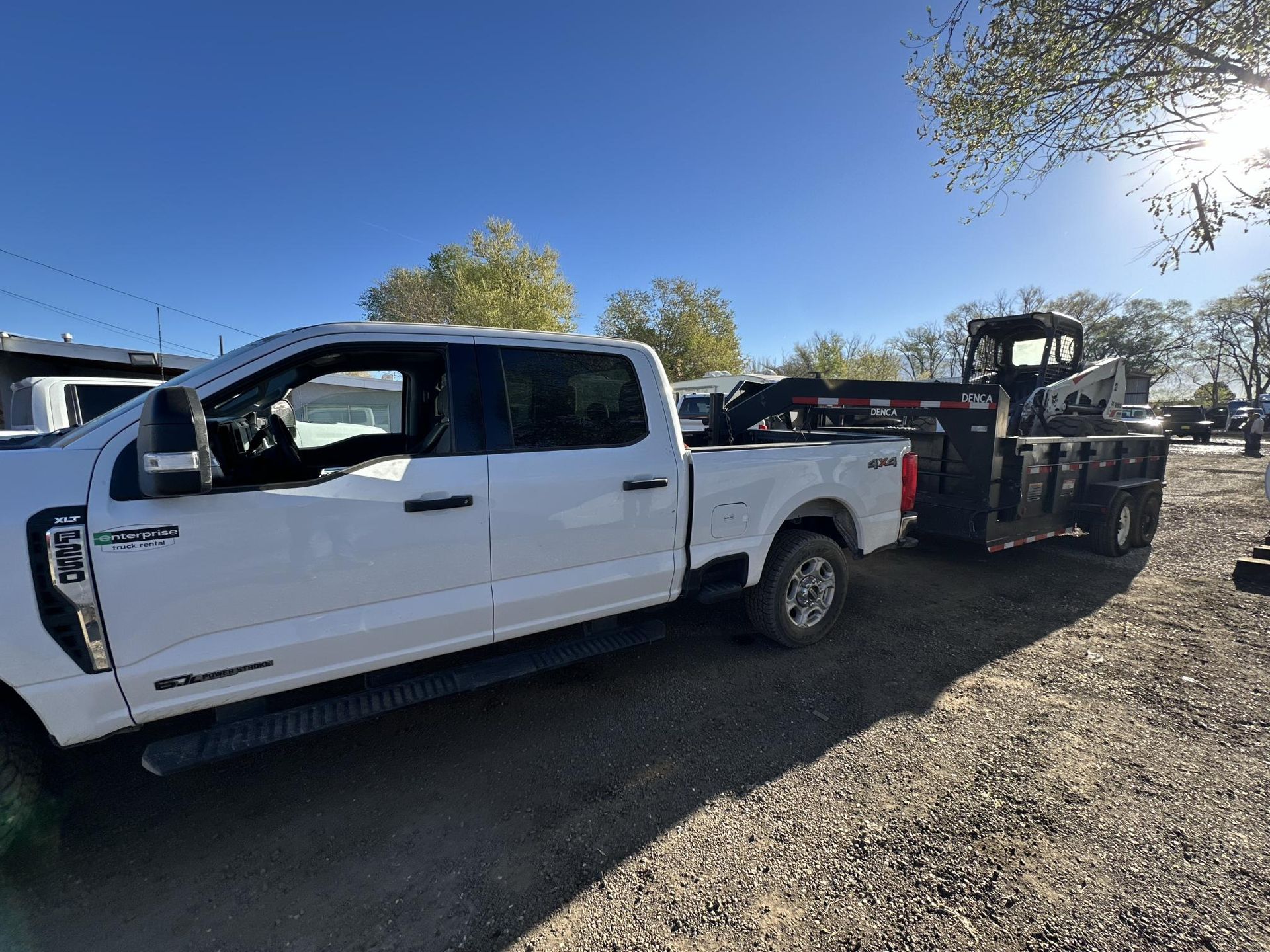 White pickup truck towing a flatbed trailer with a forklift in a gravel lot under a clear blue sky