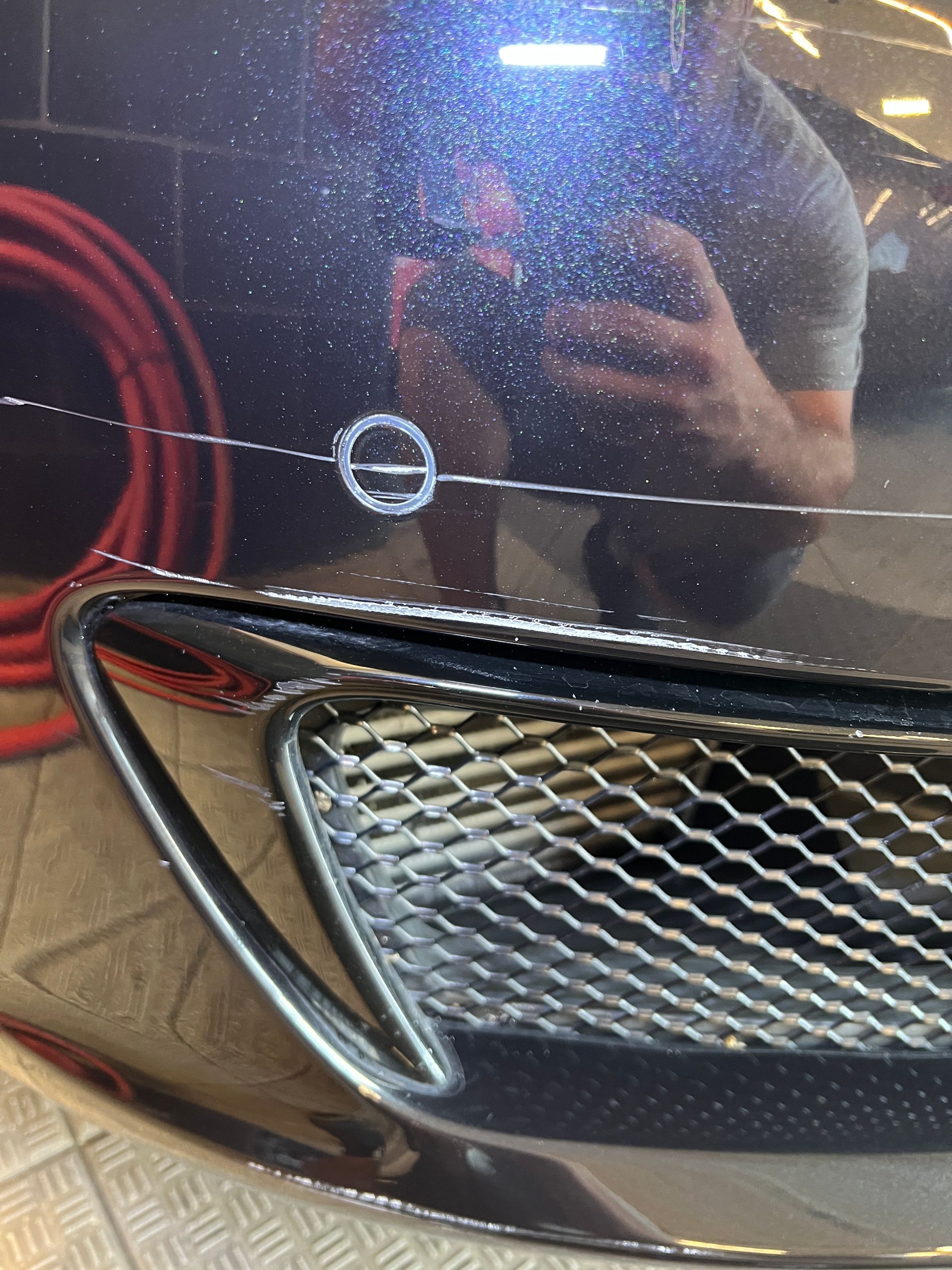 Scratched dark car bumper with a silver grill. White circular sticker visible.