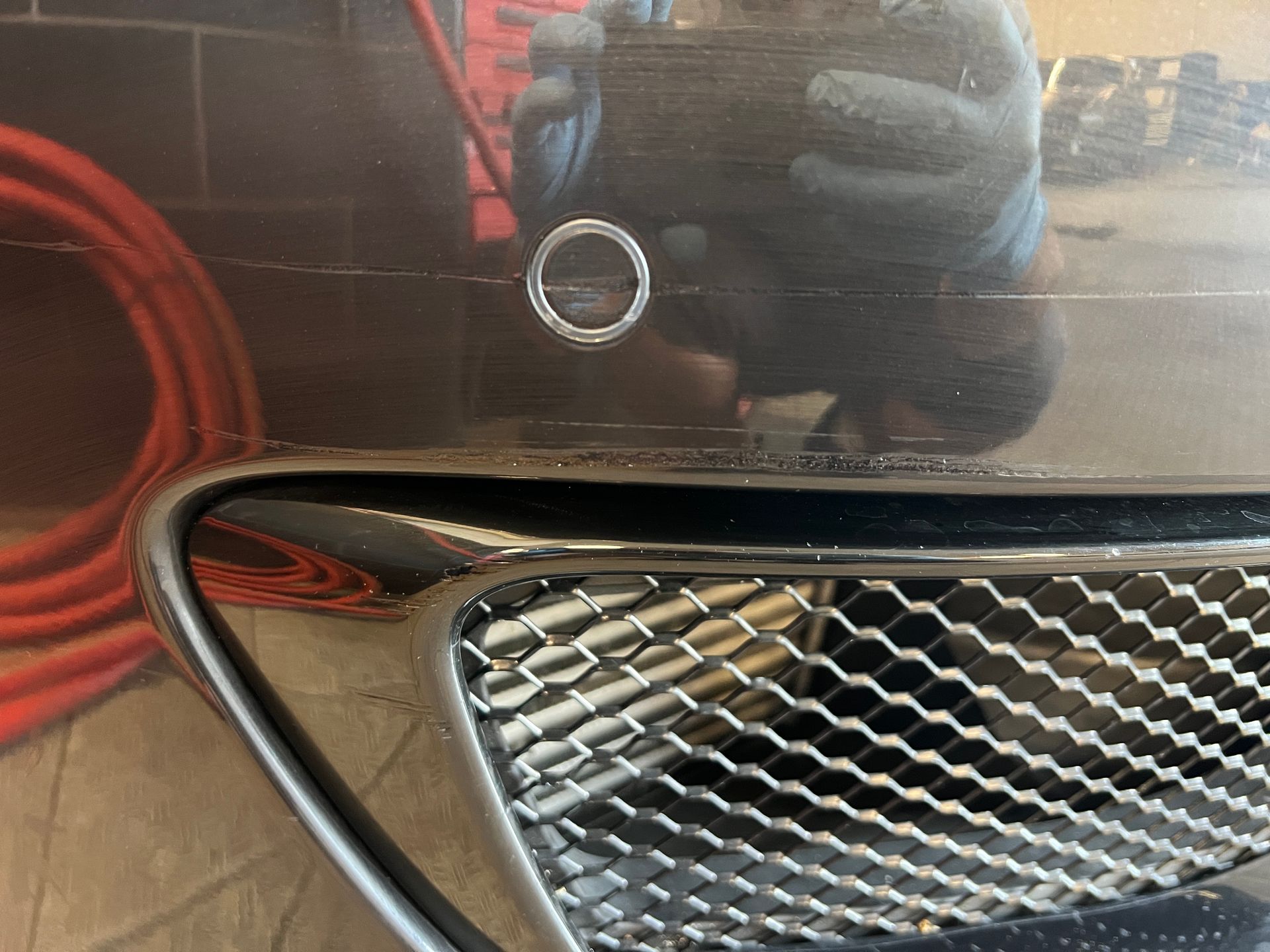 Close-up of a black car bumper with chrome ring and grill below. A person's hand is reflected in the bumper.