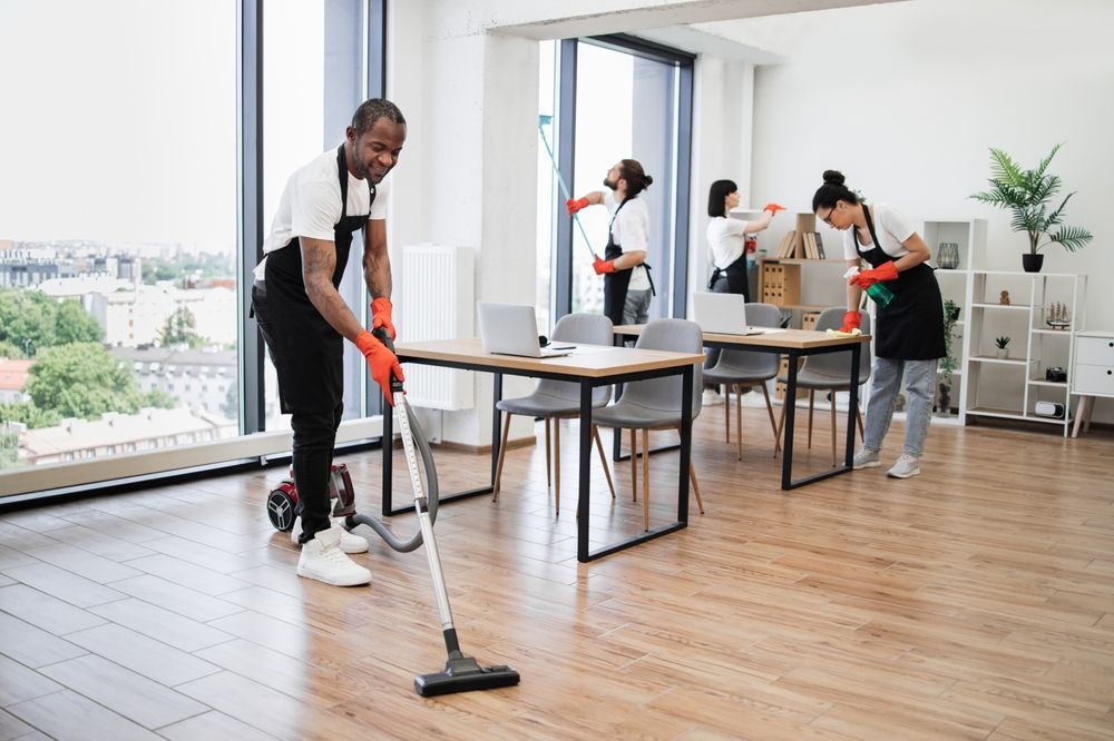 A team of four cleaning professionals in uniforms perform tasks like vacuuming and cleaning surfaces in a modern office.