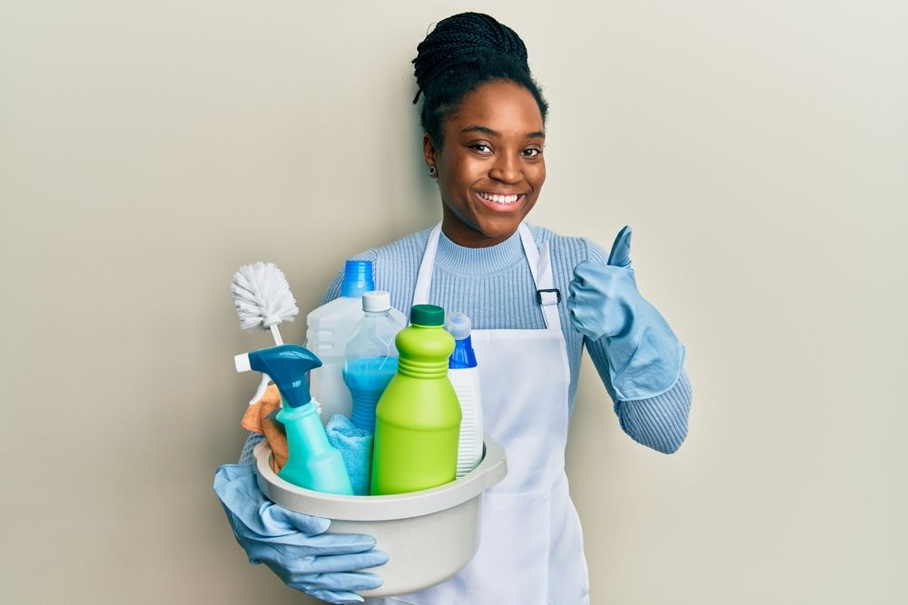Person in apron and gloves, holding cleaning supplies, giving thumbs up.