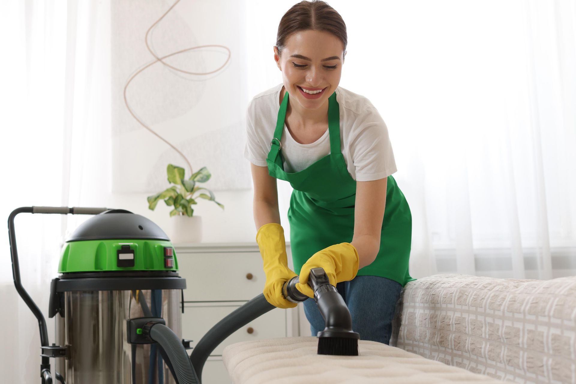 Woman in green apron vacuums a beige couch, smiling. Industrial vacuum cleaner beside her in a bright room.