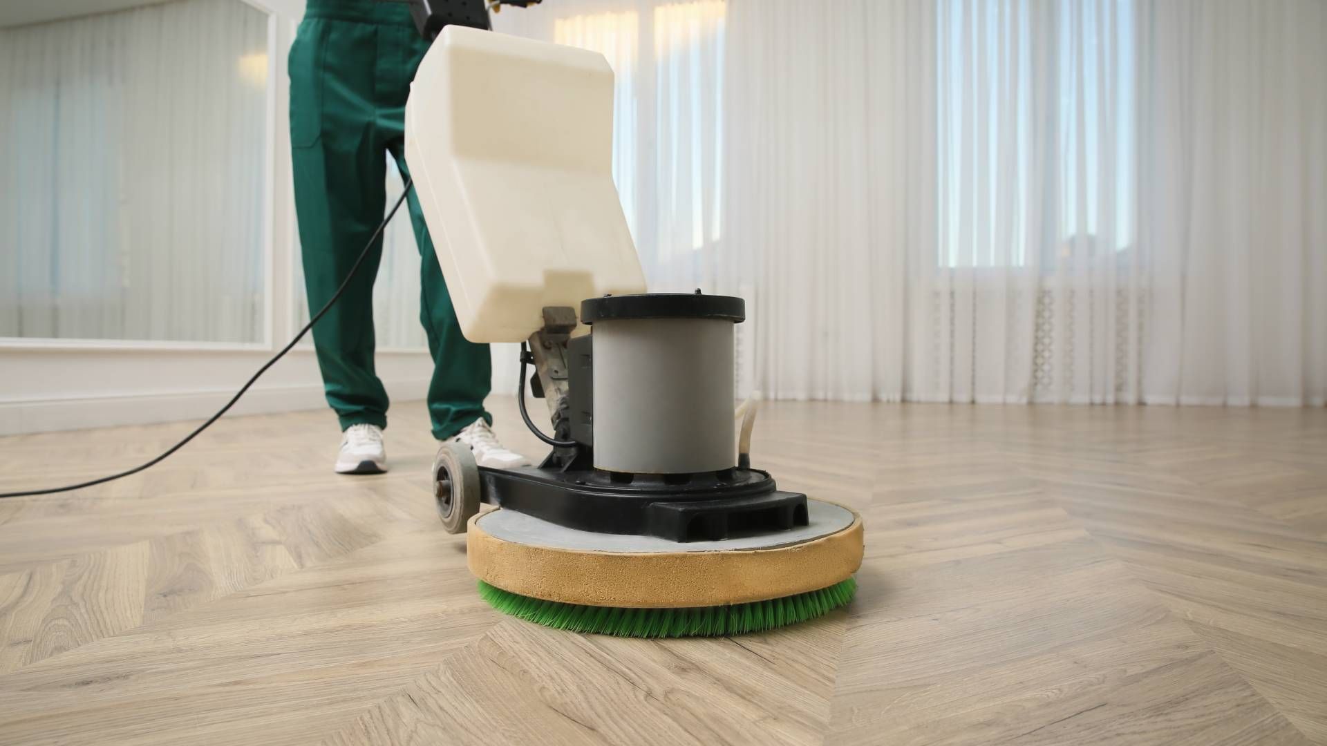 A person in green work clothing uses a floor buffer machine on a light-colored herringbone patterned wooden floor.