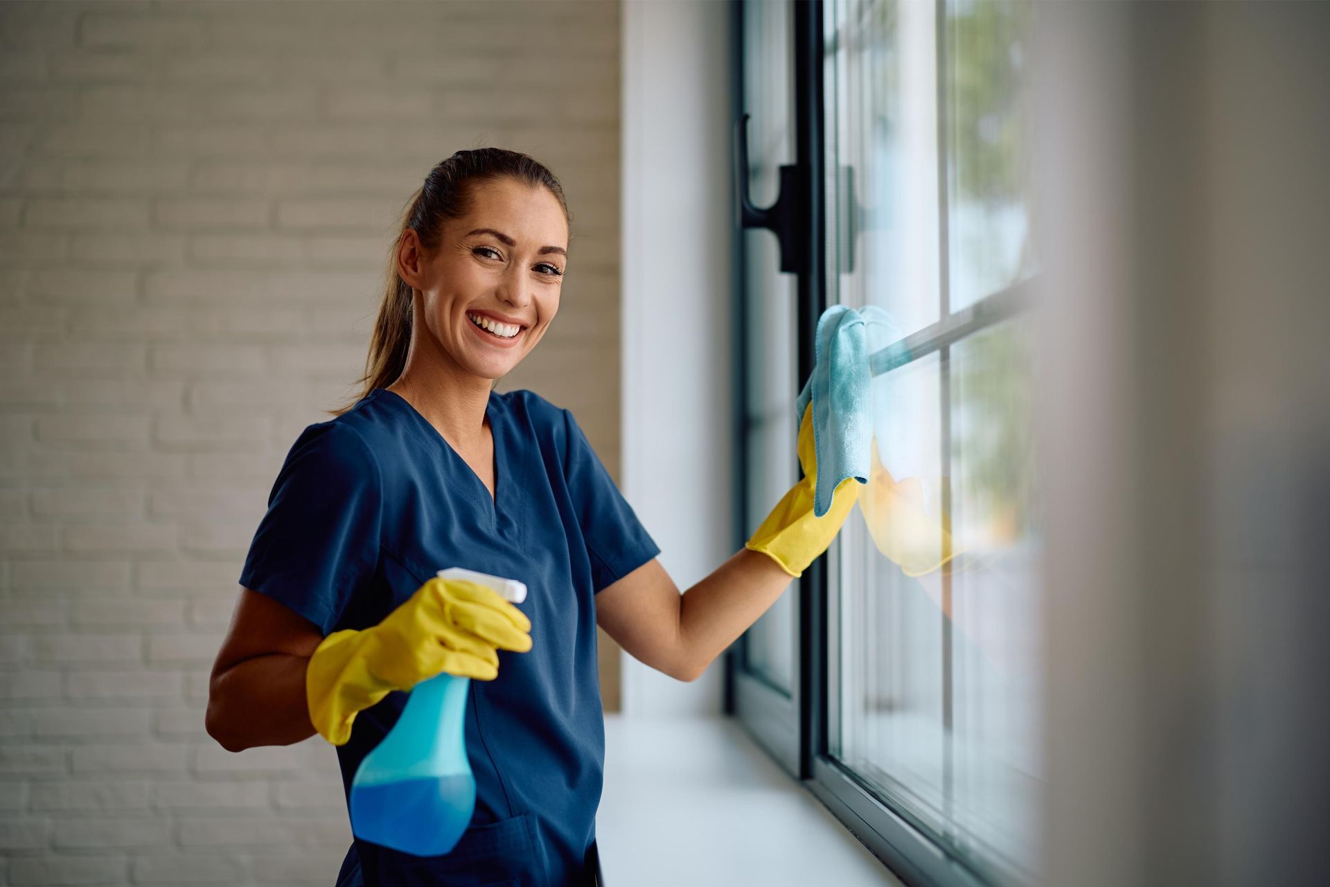 Woman wearing gloves cleaning a window with spray bottle; smiling.