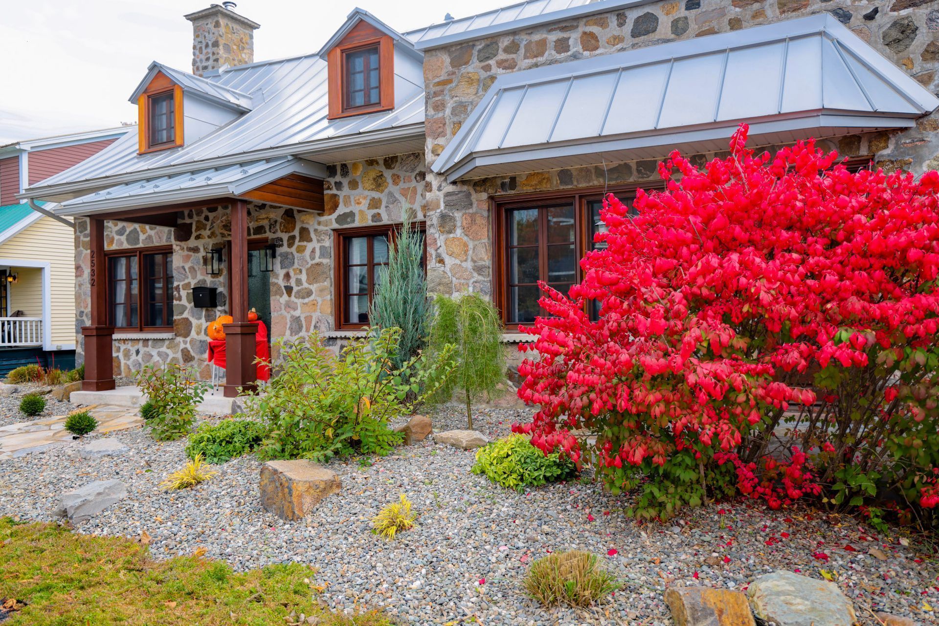 Maisonnette en pierre avec un toit en métal, des touches de rouge et un buisson fleuri rouge vif dans un jardin de gravier.