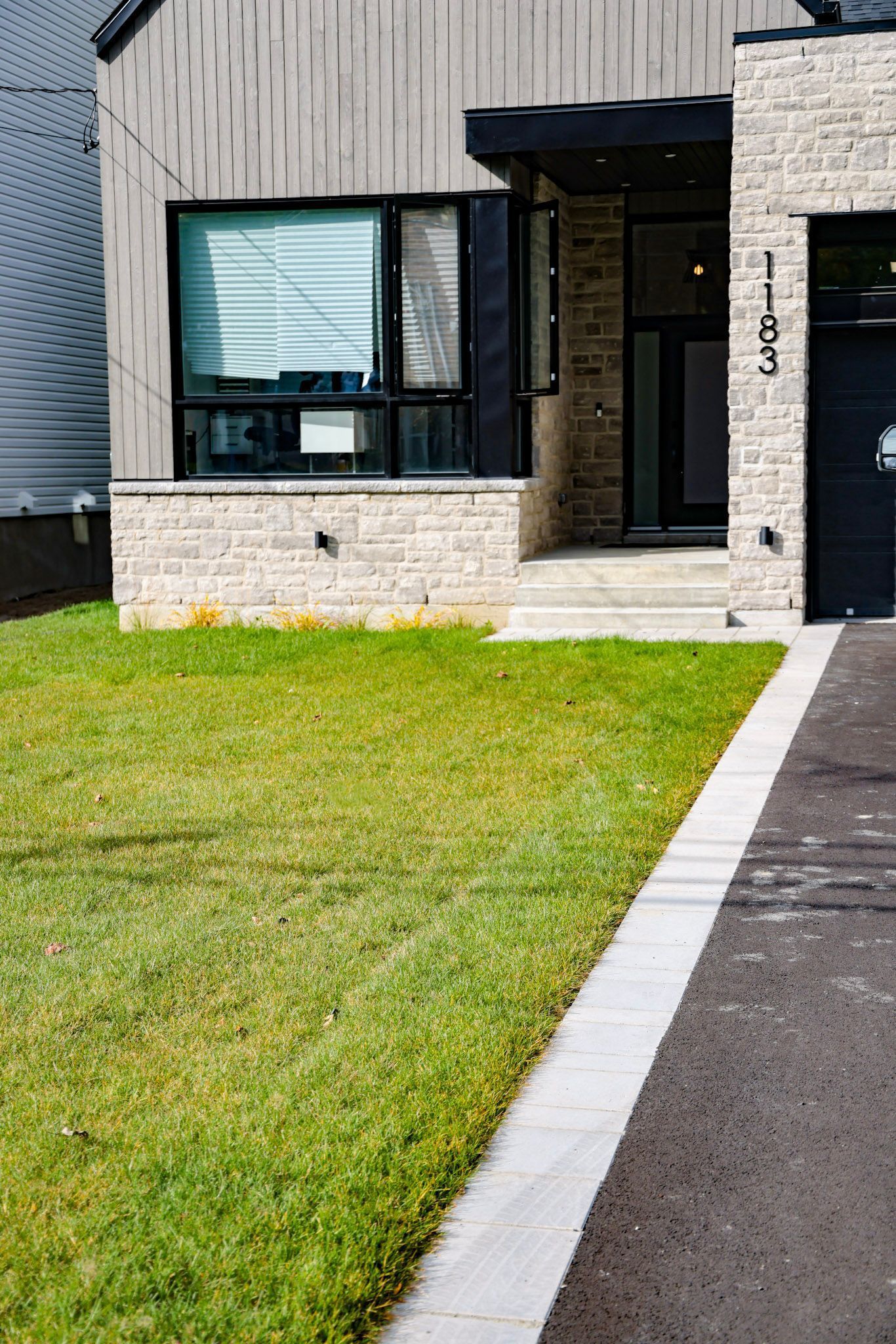 Maison moderne avec façade en pierre et bardage foncé, pelouse à l'avant et allée menant à l'entrée.