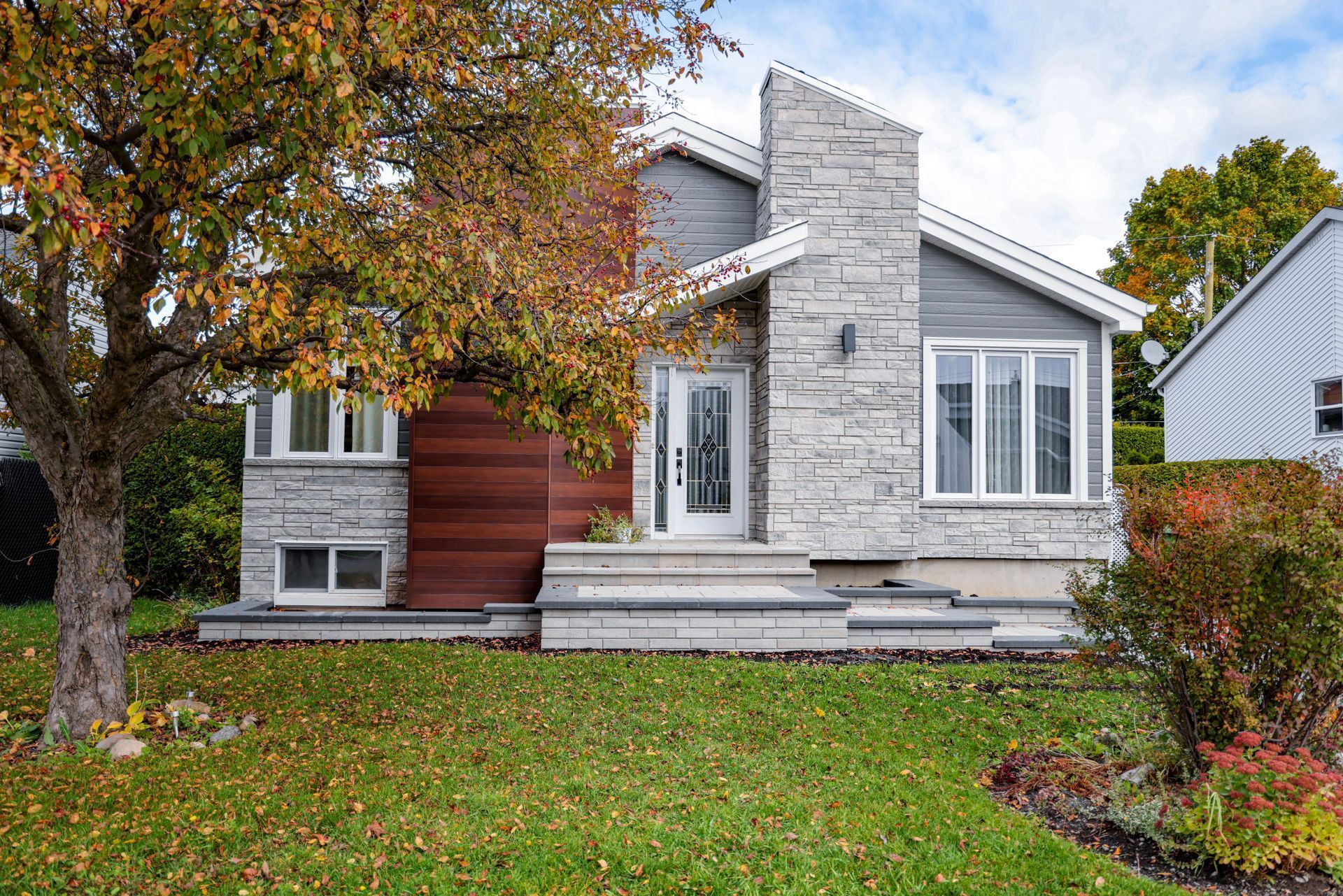 Maison moderne avec façade en pierre, porte d'entrée rouge et arbres aux couleurs d'automne dans le jardin.