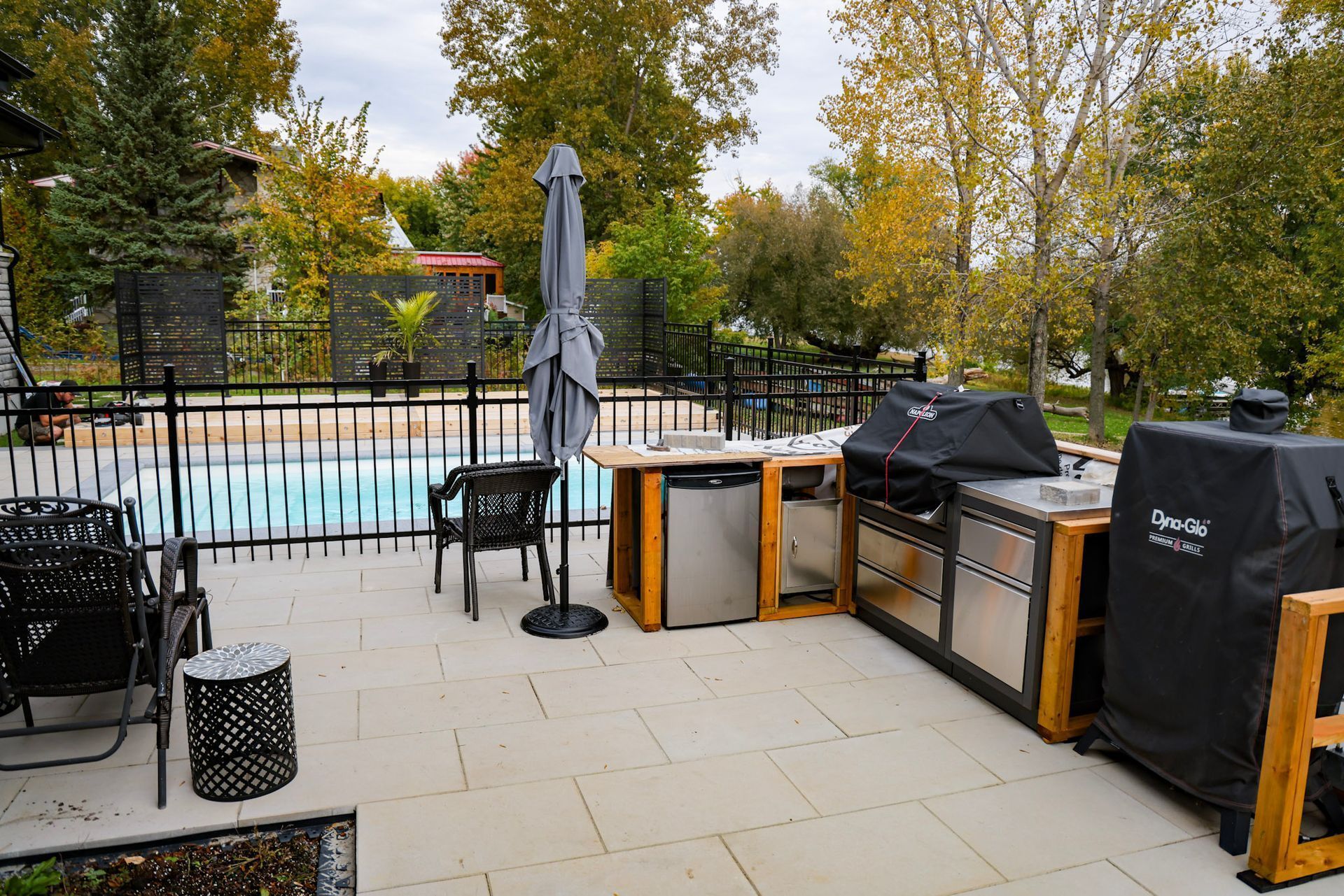 Terrasse au bord de la piscine avec parasol fermé, barbecue couvert et mobilier de jardin près d'une piscine clôturée