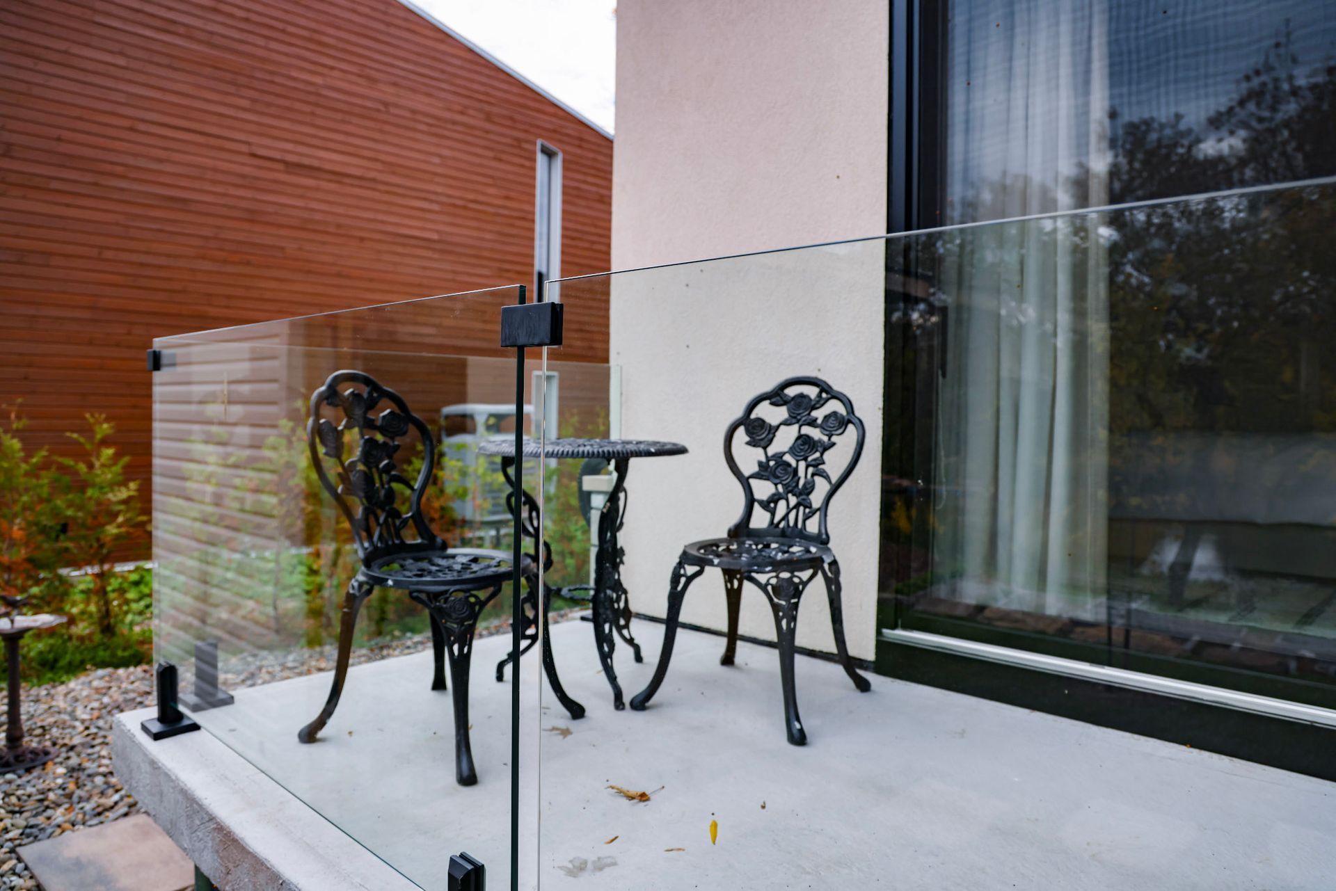 Deux chaises en fer forgé et une petite table en verre sur une terrasse moderne en béton, près d'une paroi vitrée.