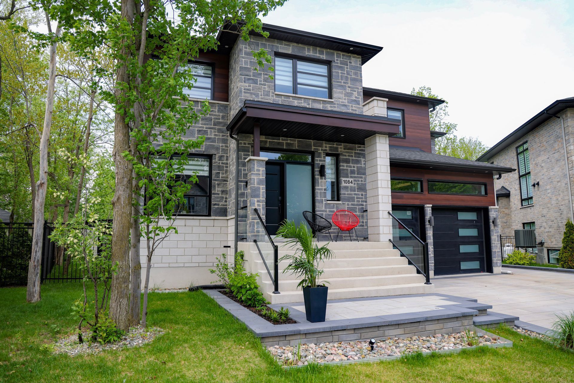 Maison moderne de deux étages avec façade en pierre, perron et garage attenant, située dans une rue résidentielle verdoyante.