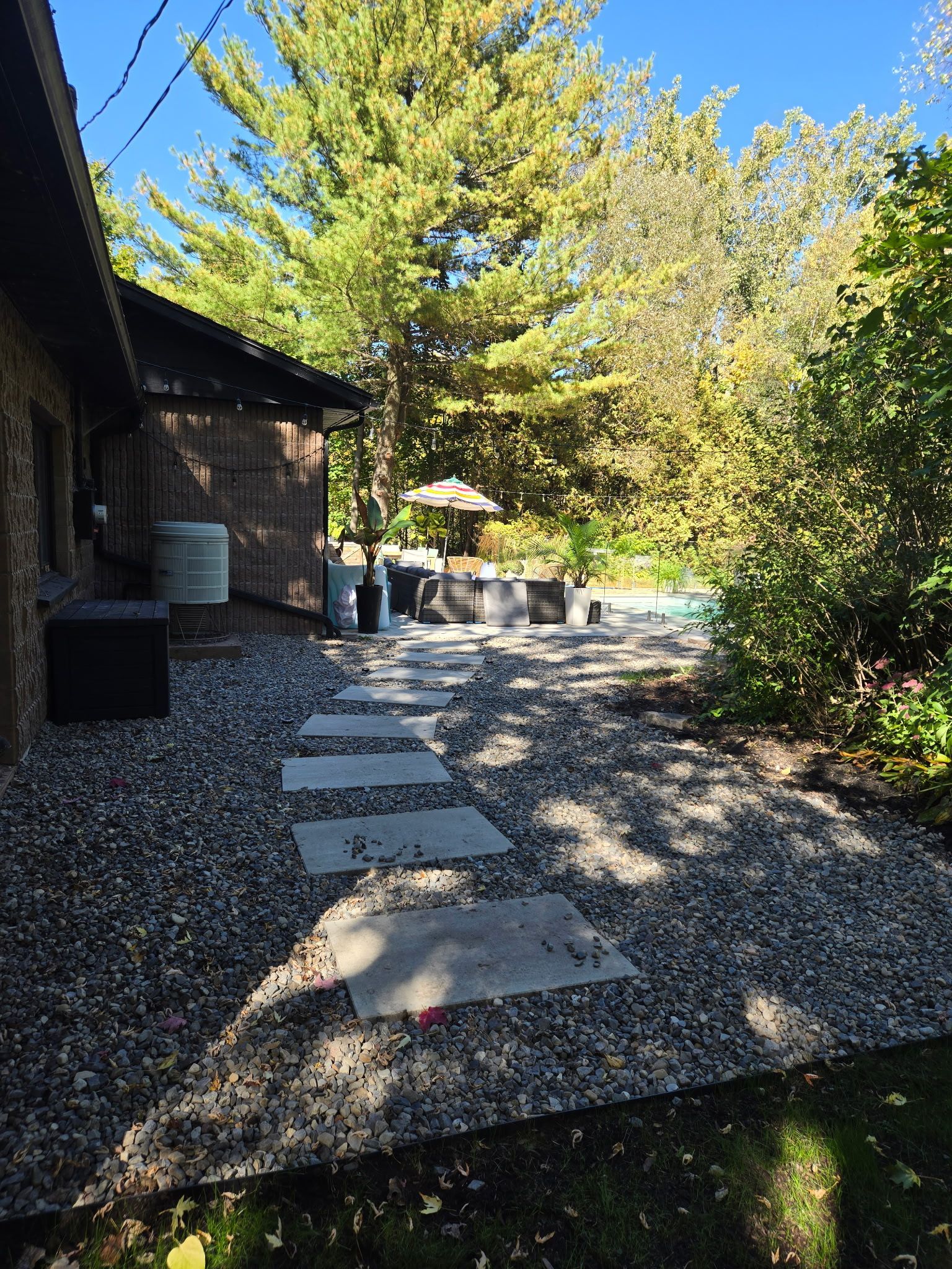 Terrasse en gravier avec pas japonais jouxtant une maison en pierre, entourée d'arbres et d'arbustes.