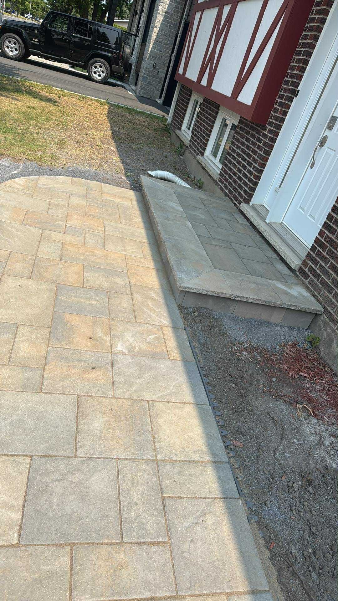Allée pavée longeant une maison avec une marche en béton, un bardage rouge et blanc et un SUV noir stationné en arrière-plan.