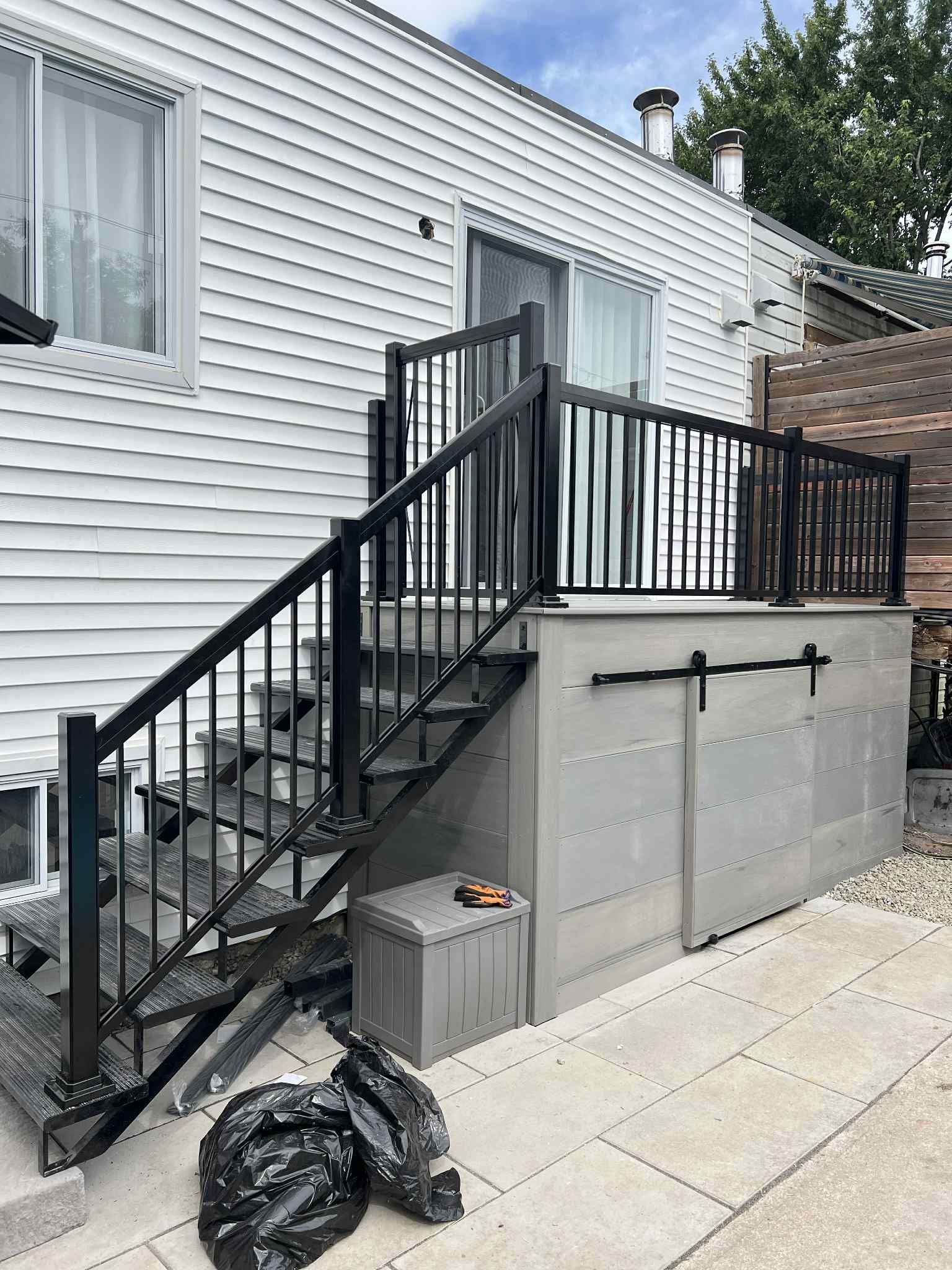 Escalier et rambarde en métal noir à côté d'une maison blanche menant à une terrasse surélevée au-dessus d'un mur de soutènement en béton.