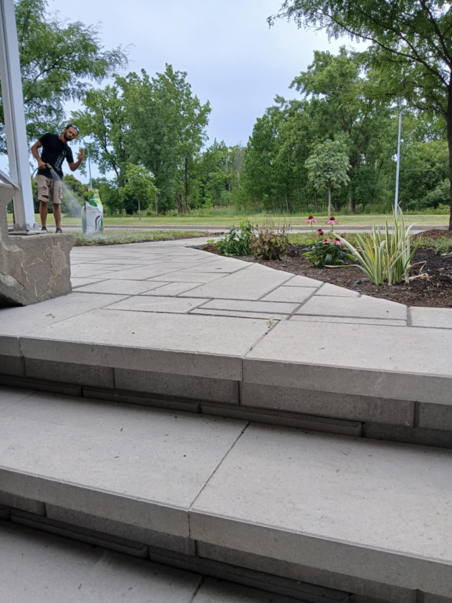 Des marches en pierre mènent à un chemin de jardin pavé bordé d'arbres et de parterres de fleurs ; deux personnes se trouvent à gauche.