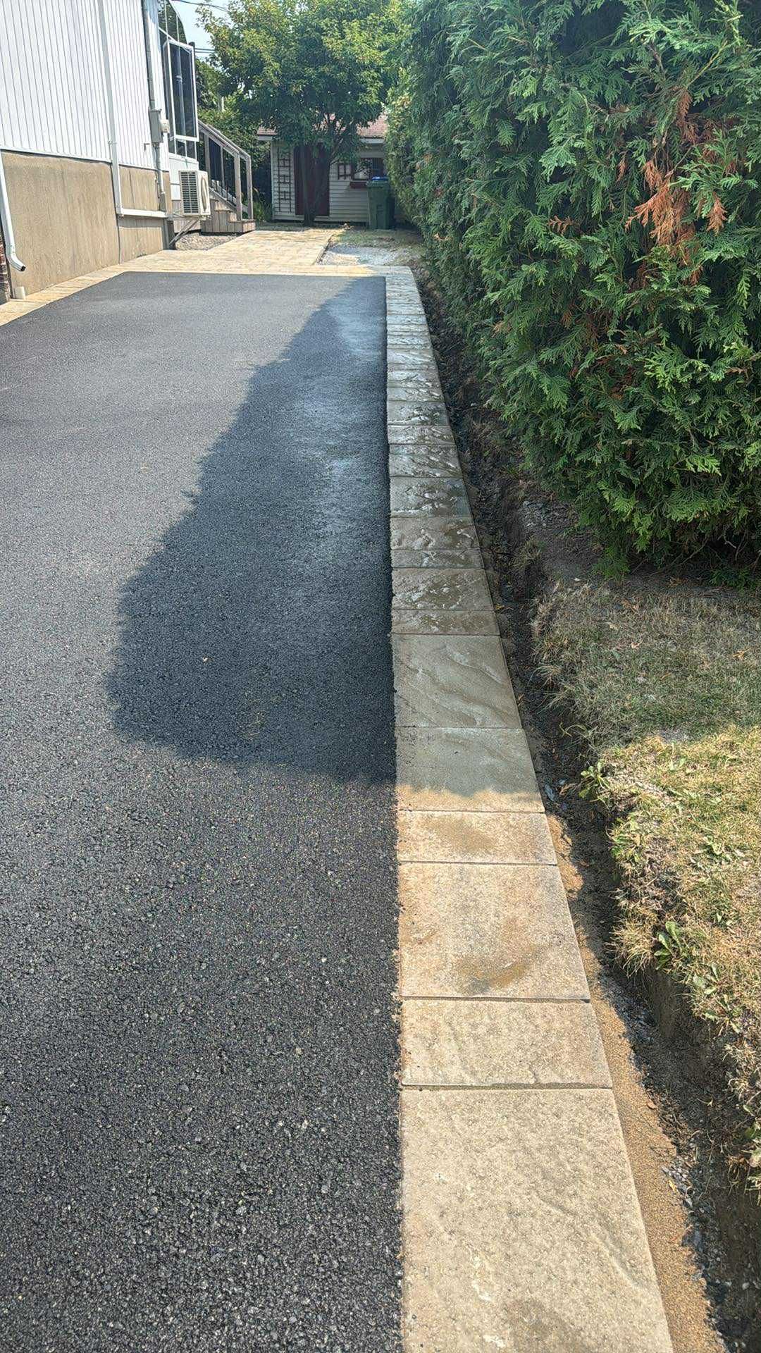 Allée pavée étroite, bordée d'une haie et d'un trottoir en pierre, menant à un portail baigné de soleil.