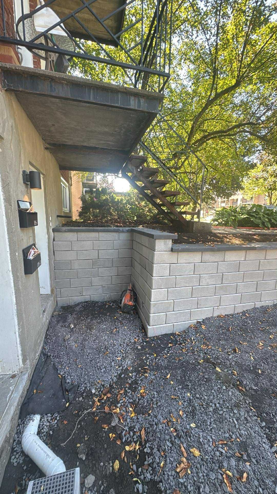 Terrasse extérieure en béton avec un muret en parpaings, sol en gravier et façade de maison sous les arbres.