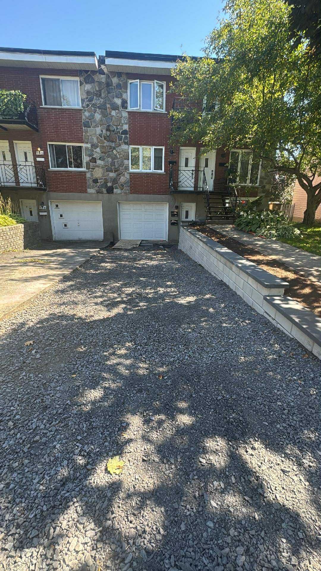 Maison de ville en briques avec deux portes de garage, allée en gravier et arbres projetant des ombres à l'avant.
