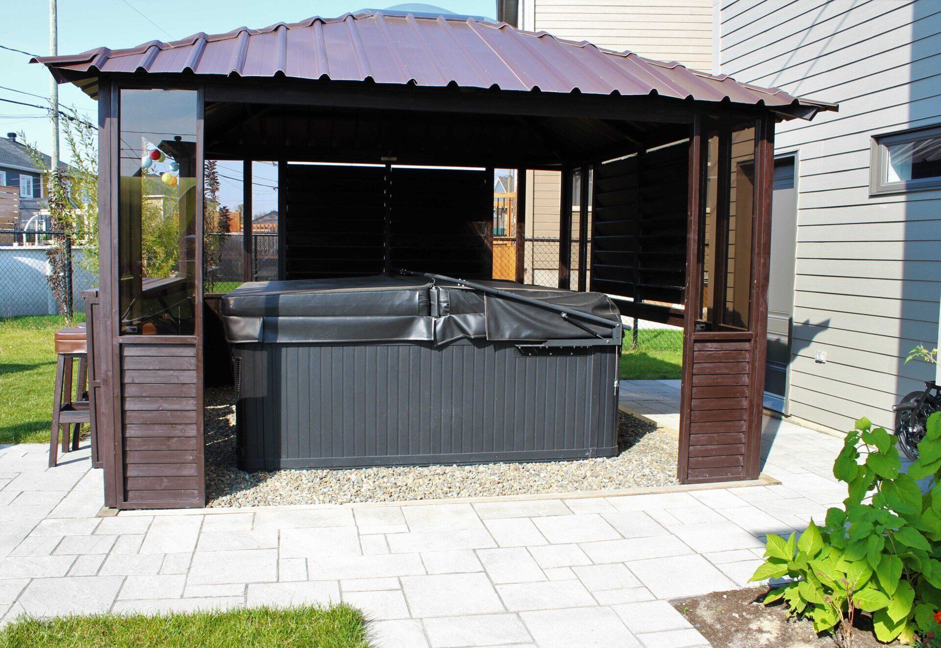 Espace jacuzzi extérieur couvert avec un gazebo marron foncé et un spa noir dans un jardin.