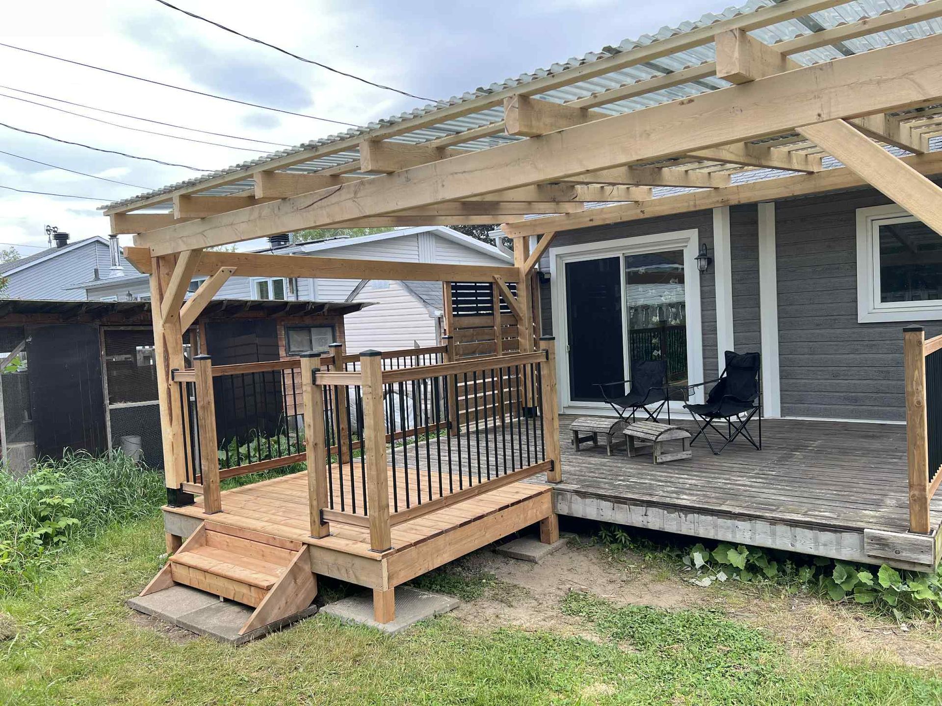 Petit porche en bois et rampe attenants à une maison grise, avec des chaises de patio et une pergola au-dessus.