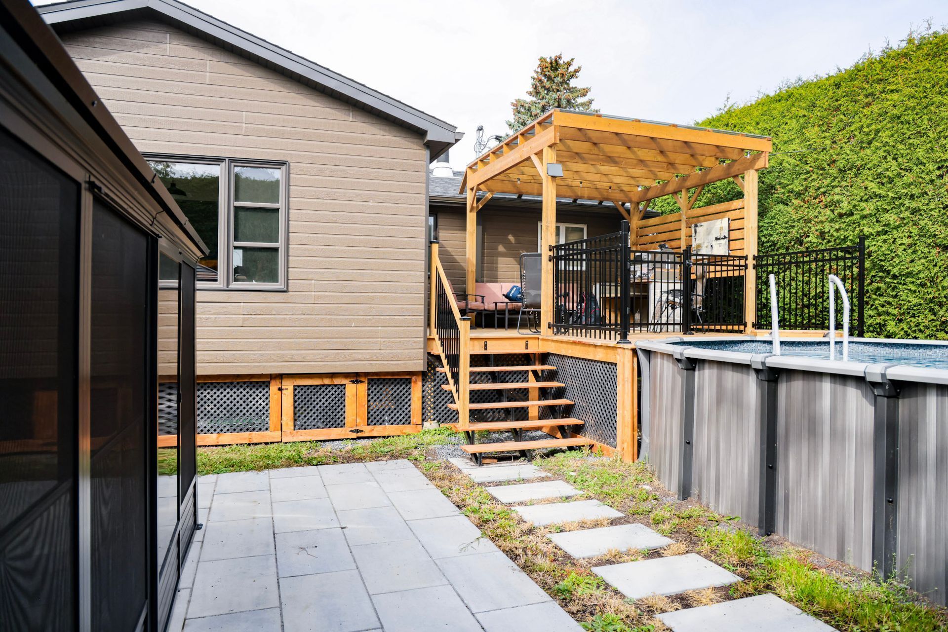 Terrasse arrière avec pavés gris, escalier menant à une pergola en bois et piscine hors sol à côté de la maison