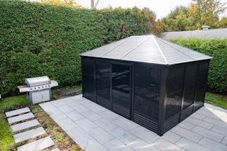 Terrasse arrière avec un gazebo noir et un barbecue en acier inoxydable à côté d'une haute haie.