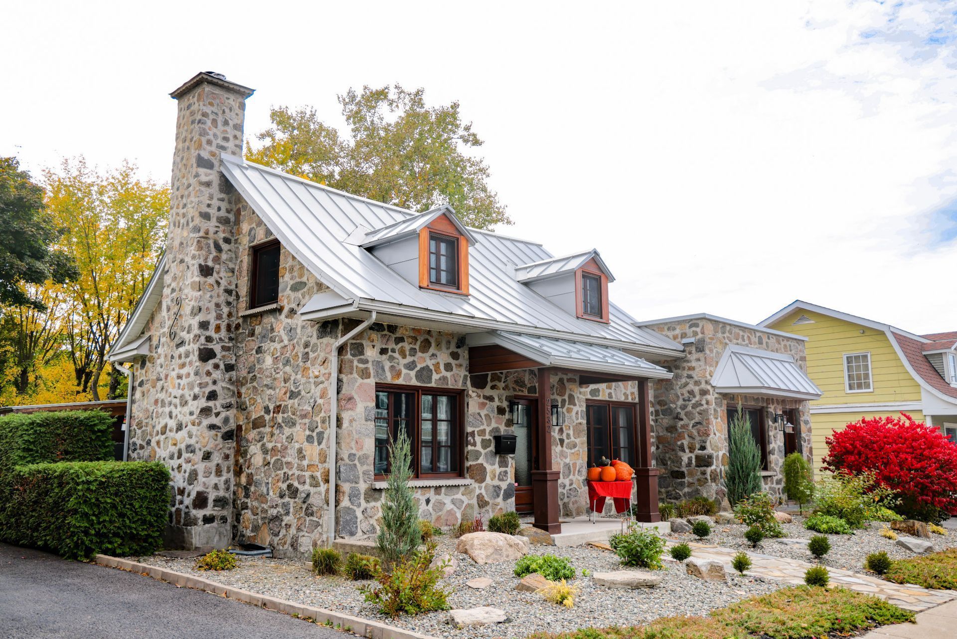 Maisonnette en pierre avec toit en tôle, jardin aux couleurs d'automne, et une personne en veste orange sur le porche