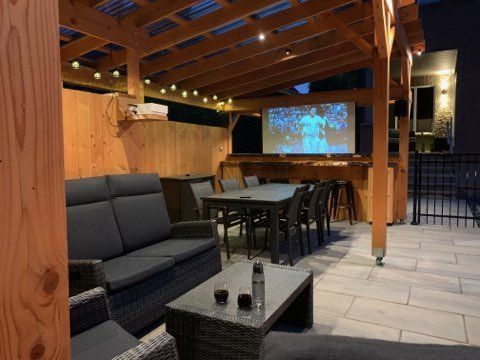 Salon extérieur couvert avec sièges sombres, tables, guirlandes lumineuses et écran de projection sous un toit en bois.