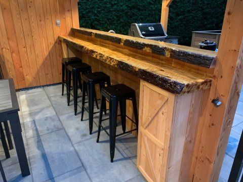 Bar extérieur rustique avec comptoir en bois, tabourets noirs et ordinateur portable posé dessus.
