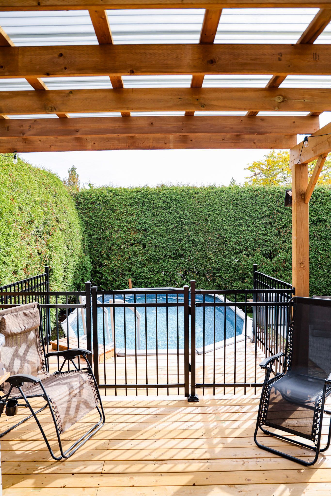 Terrasse couverte avec pergola en bois, chaises longues et piscine clôturée dans le jardin.